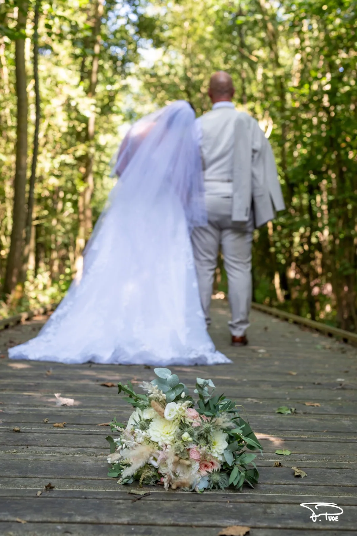 Les mariés marchent de dos sur un chemin en bois dans la forêt, le bouquet au premier plan