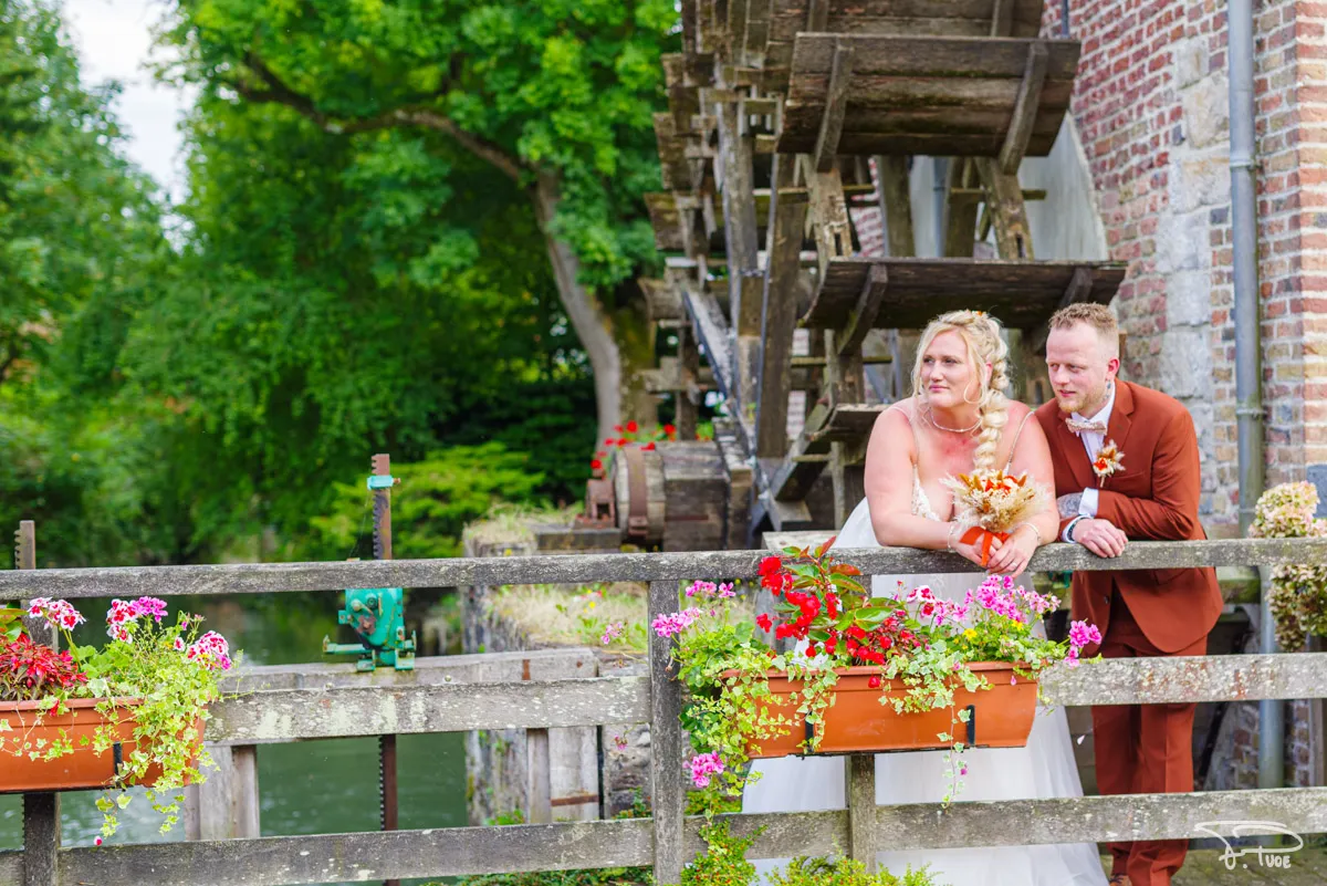 Les mariés accoudés à une passerelle fleurie devant la roue d’un moulin