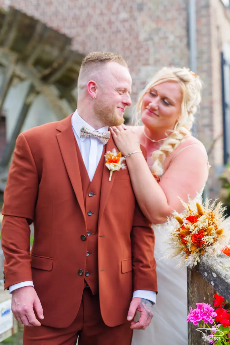 La mariée pose la main sur l’épaule du marié en costume rouille, devant un moulin