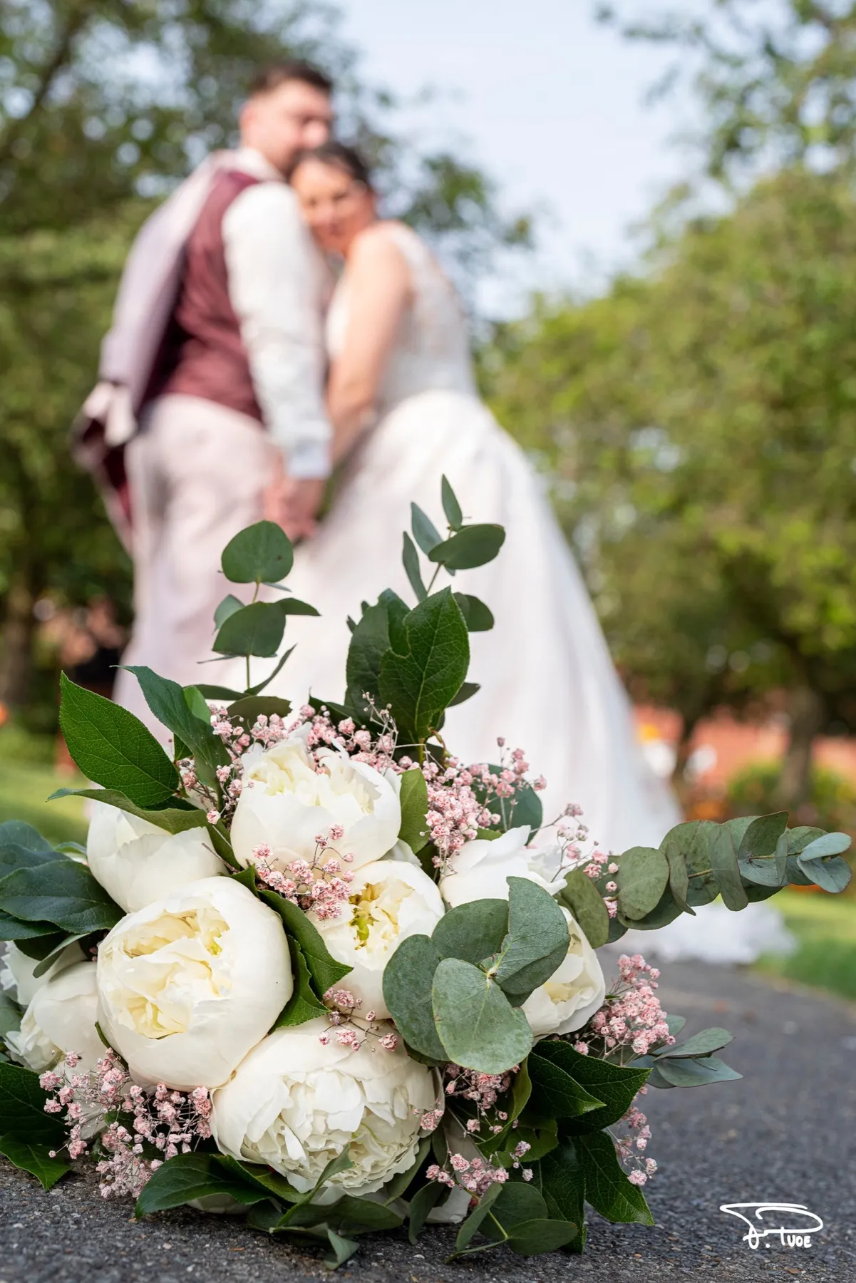 Un bouquet de pivoines blanches au premier plan, les mariés enlacés en arrière-plan