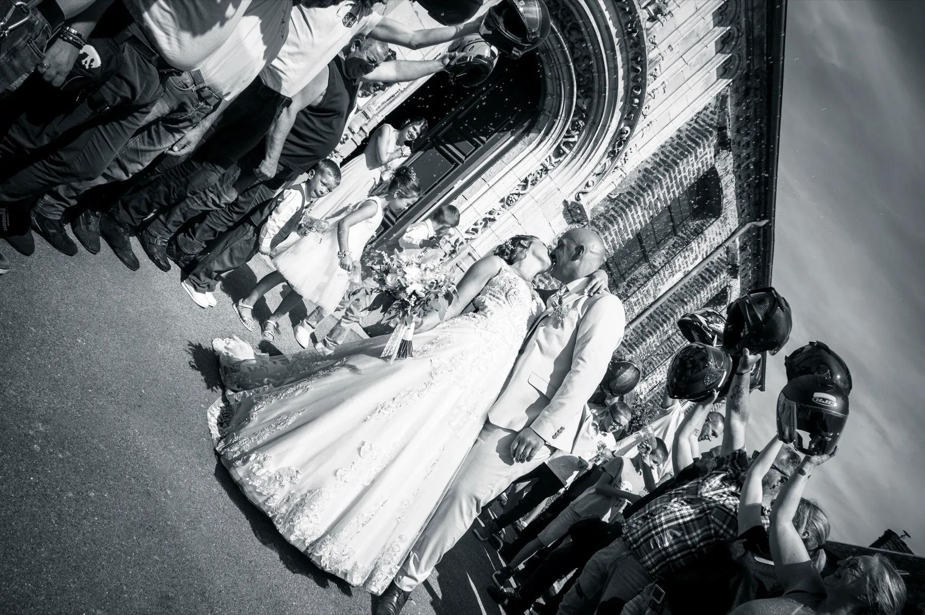 Les mariés s’embrassent à la sortie de l’église, entourés des enfants d’honneur et des invités, en noir et blanc