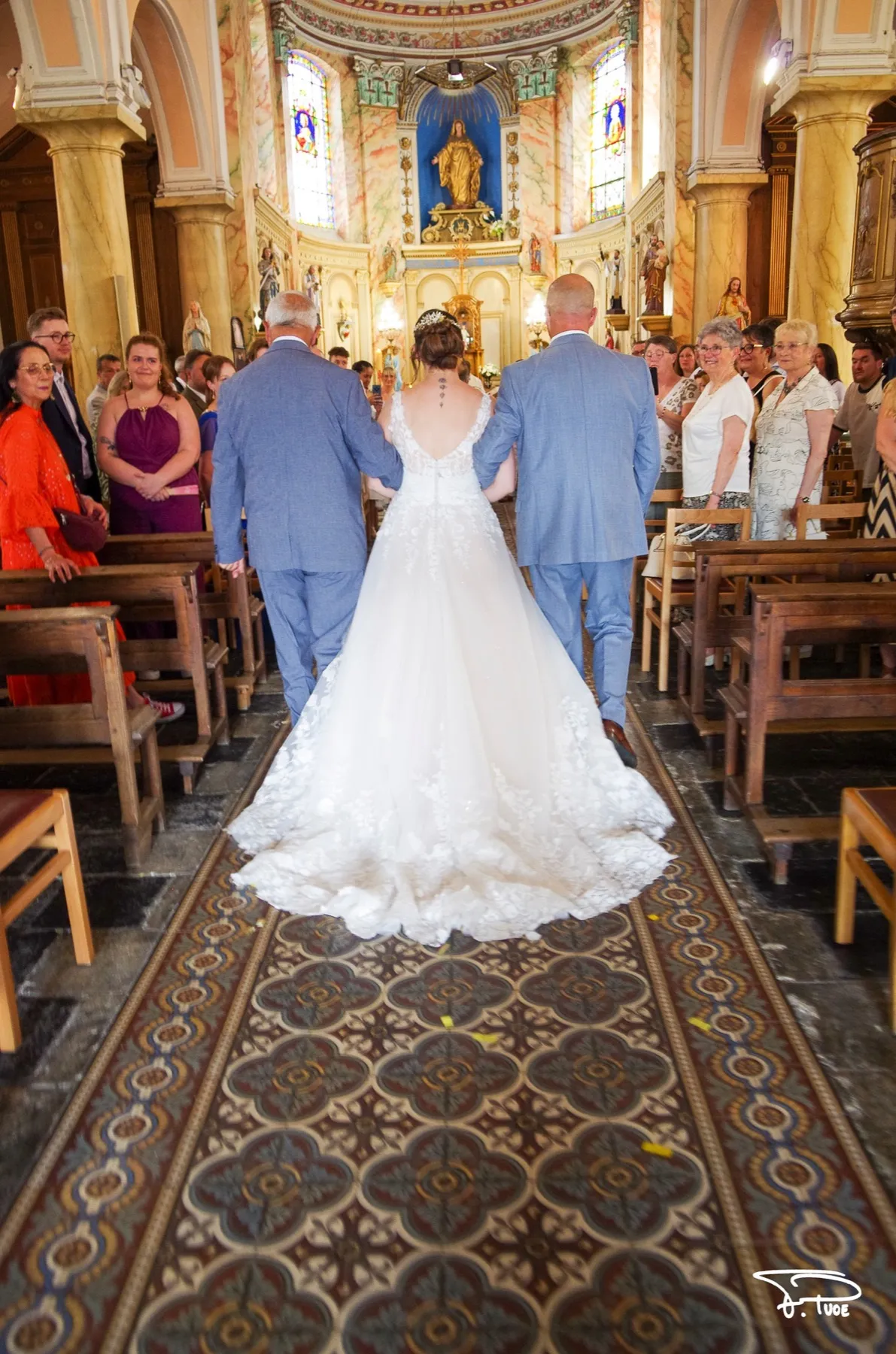 Vue de dos, la mariée remonte l’allée de l’église entre deux hommes en costume bleu, sous le regard des invités