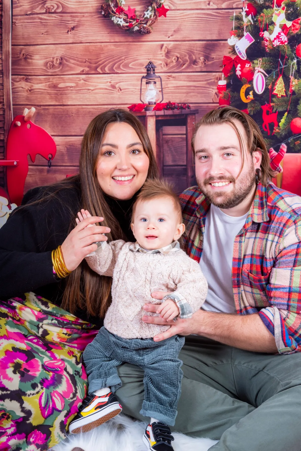 Jeunes parents souriants avec leur bébé devant un décor de chalet et de sapin de Noël en studio