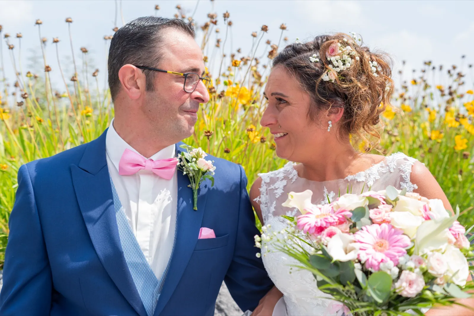 Le marié et la mariée se regardent en souriant devant des fleurs jaunes sous un ciel clair