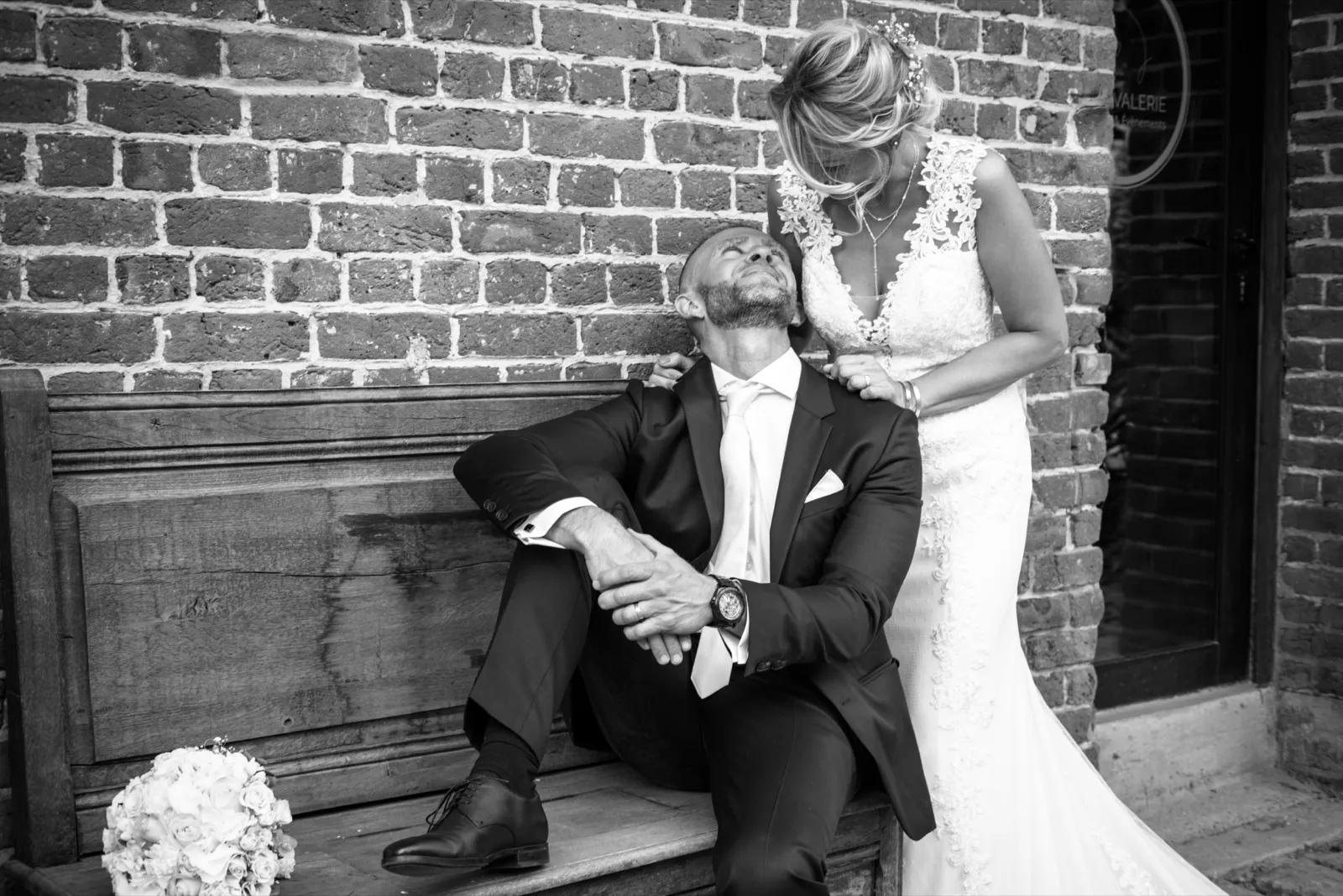 En noir et blanc, la mariée tient les épaules du marié qui renverse la tête en riant sur un banc