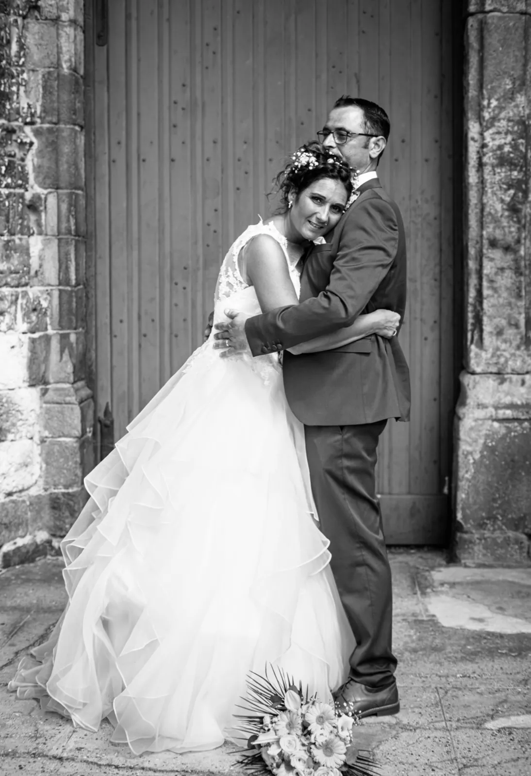 En noir et blanc, les mariés s'enlacent tendrement devant une grande porte en bois ancienne