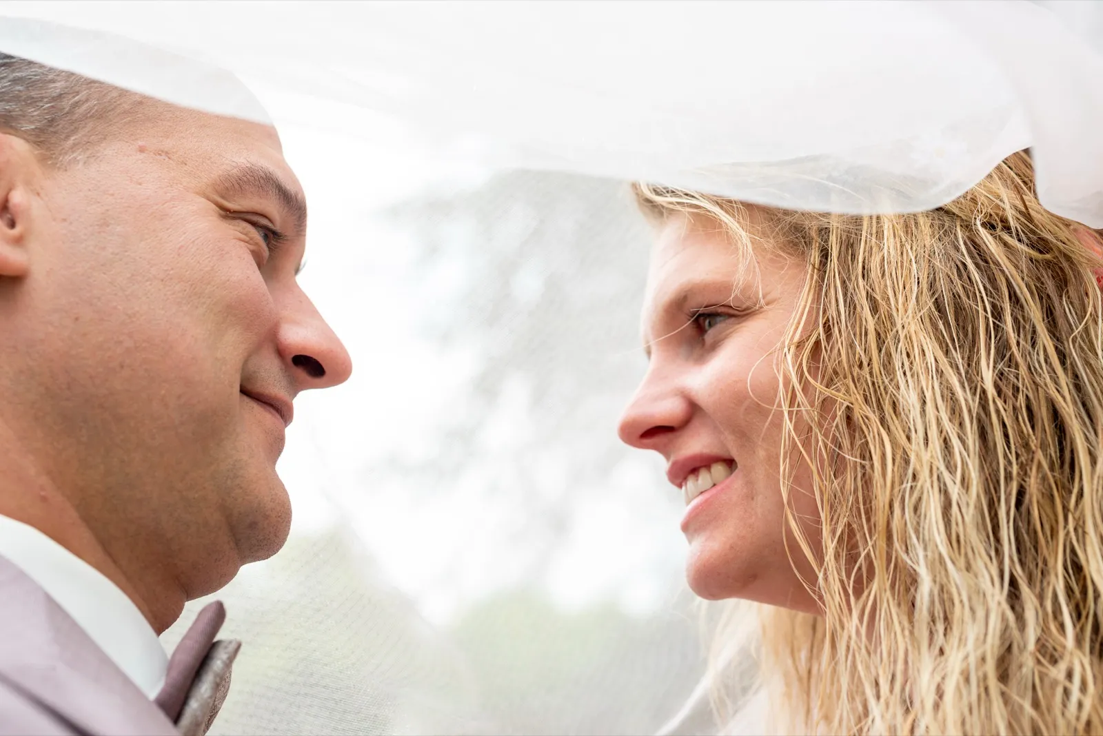 Sous un voile blanc, les mariés se regardent en souriant, visages tout proches