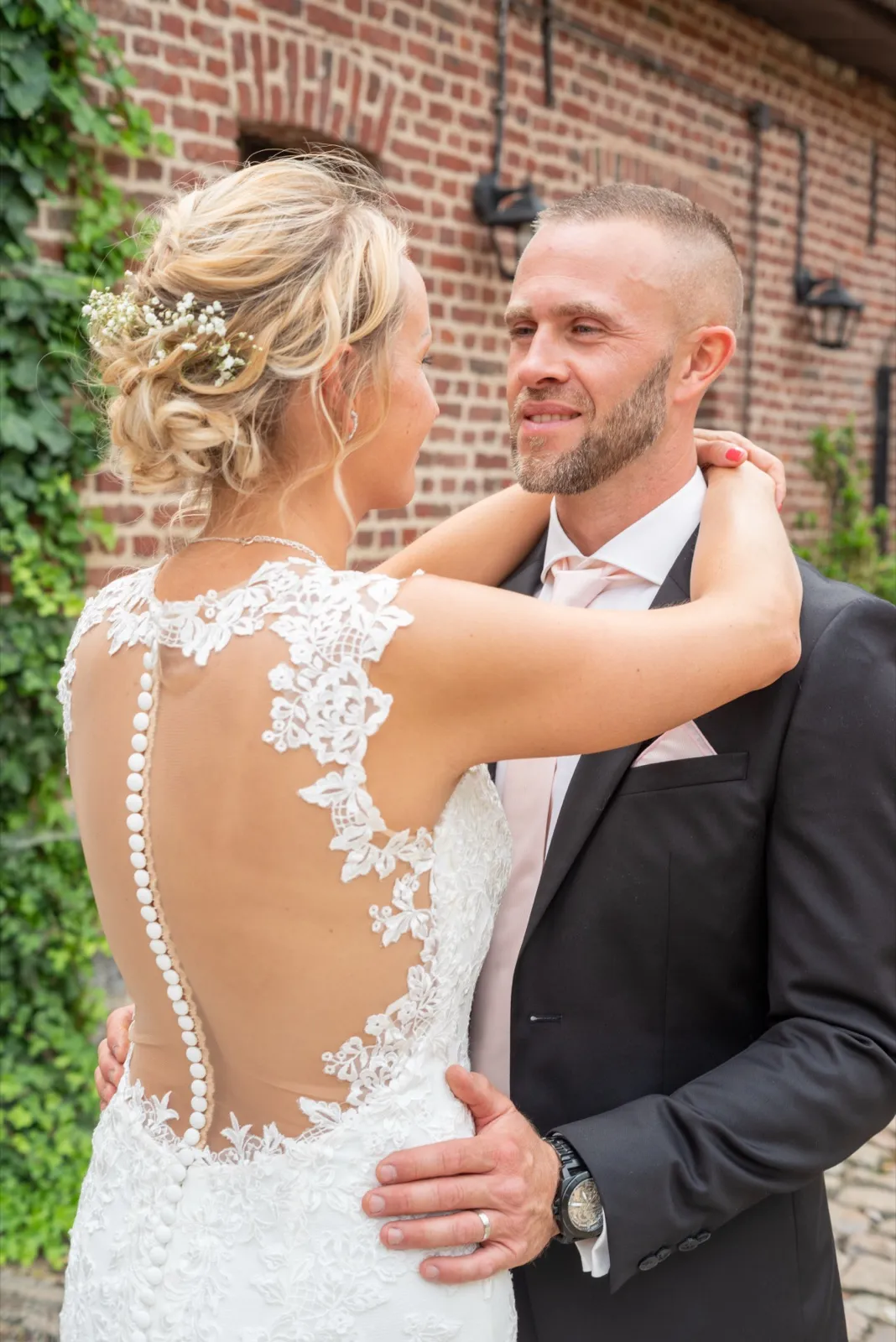 Dos en dentelle boutonné, la mariée enlace le marié qui lui sourit devant un mur de briques