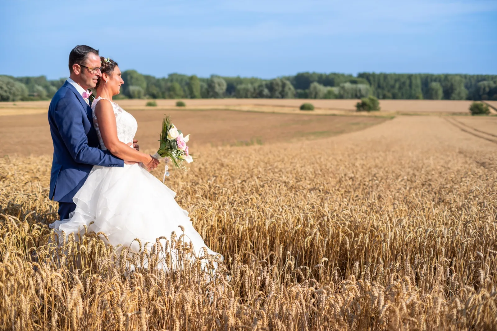 Les mariés enlacés regardent au loin, debout dans un champ de blé sous le ciel bleu