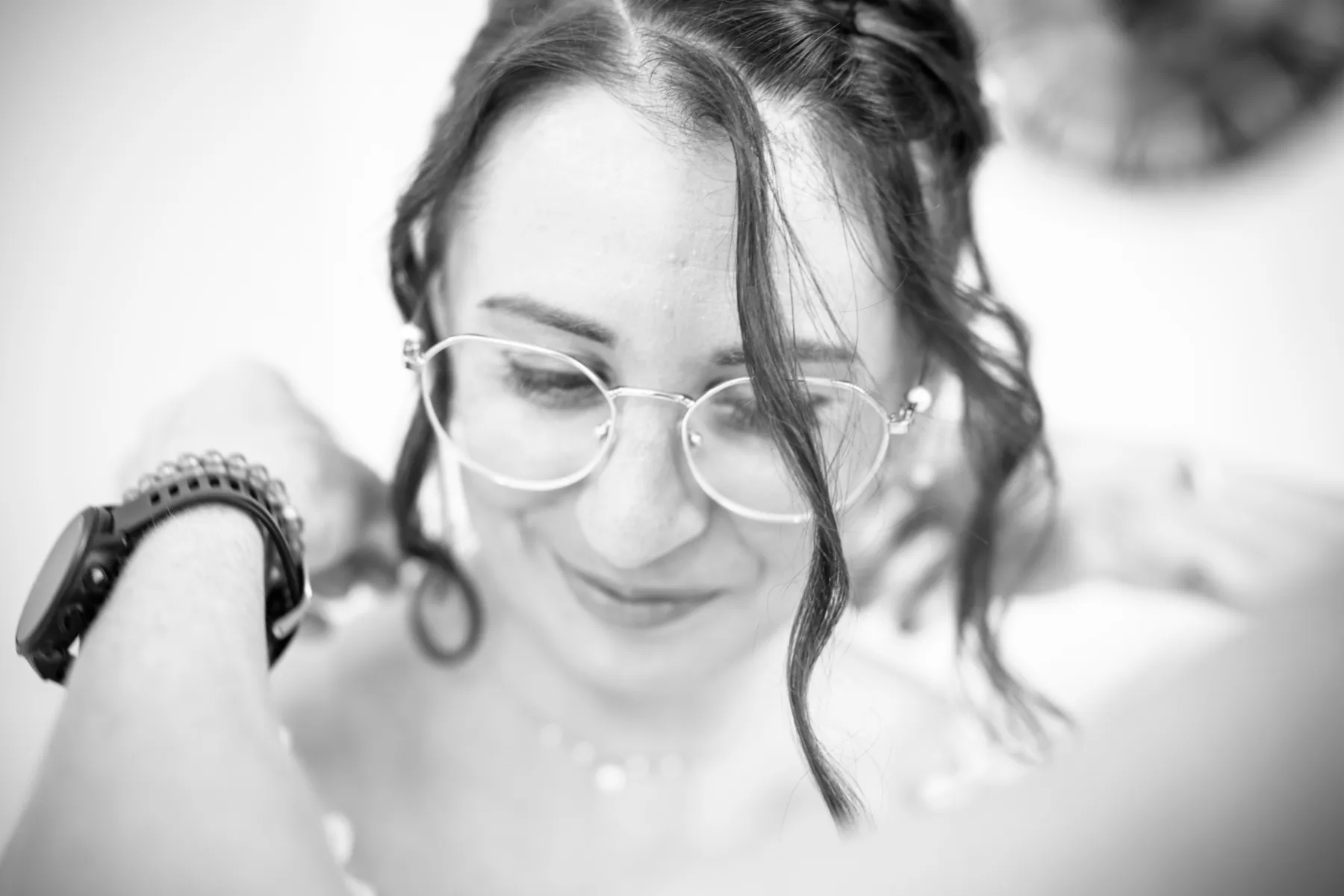 En noir et blanc, la mariée sourit, les yeux baissés, les mains portées à sa nuque
