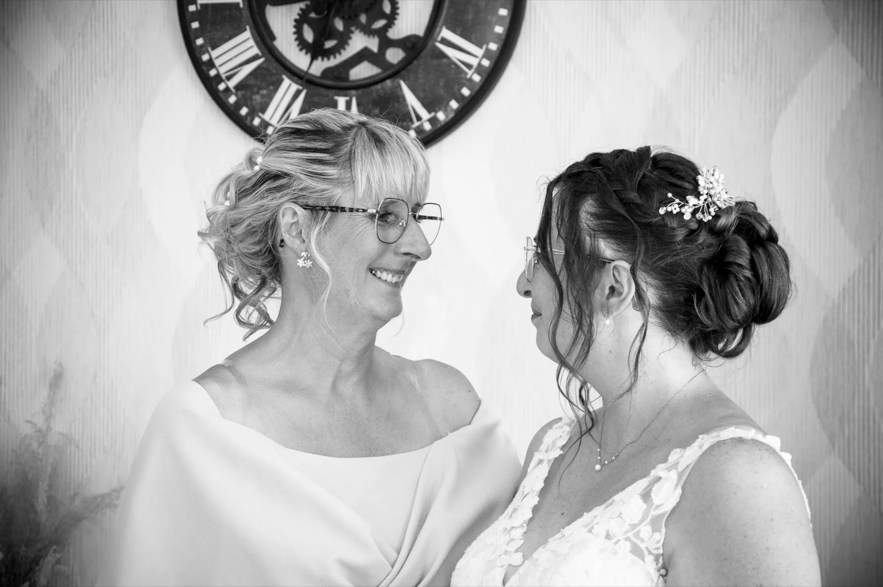 En noir et blanc, la mariée face à une proche souriante, sous une grande horloge