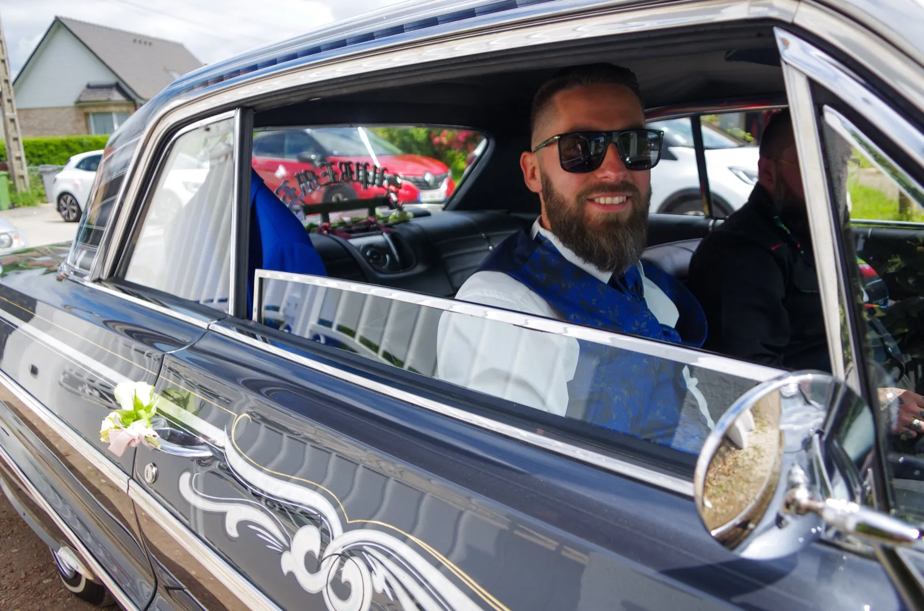 Le marié, lunettes de soleil et large sourire, à la fenêtre d’une voiture ancienne