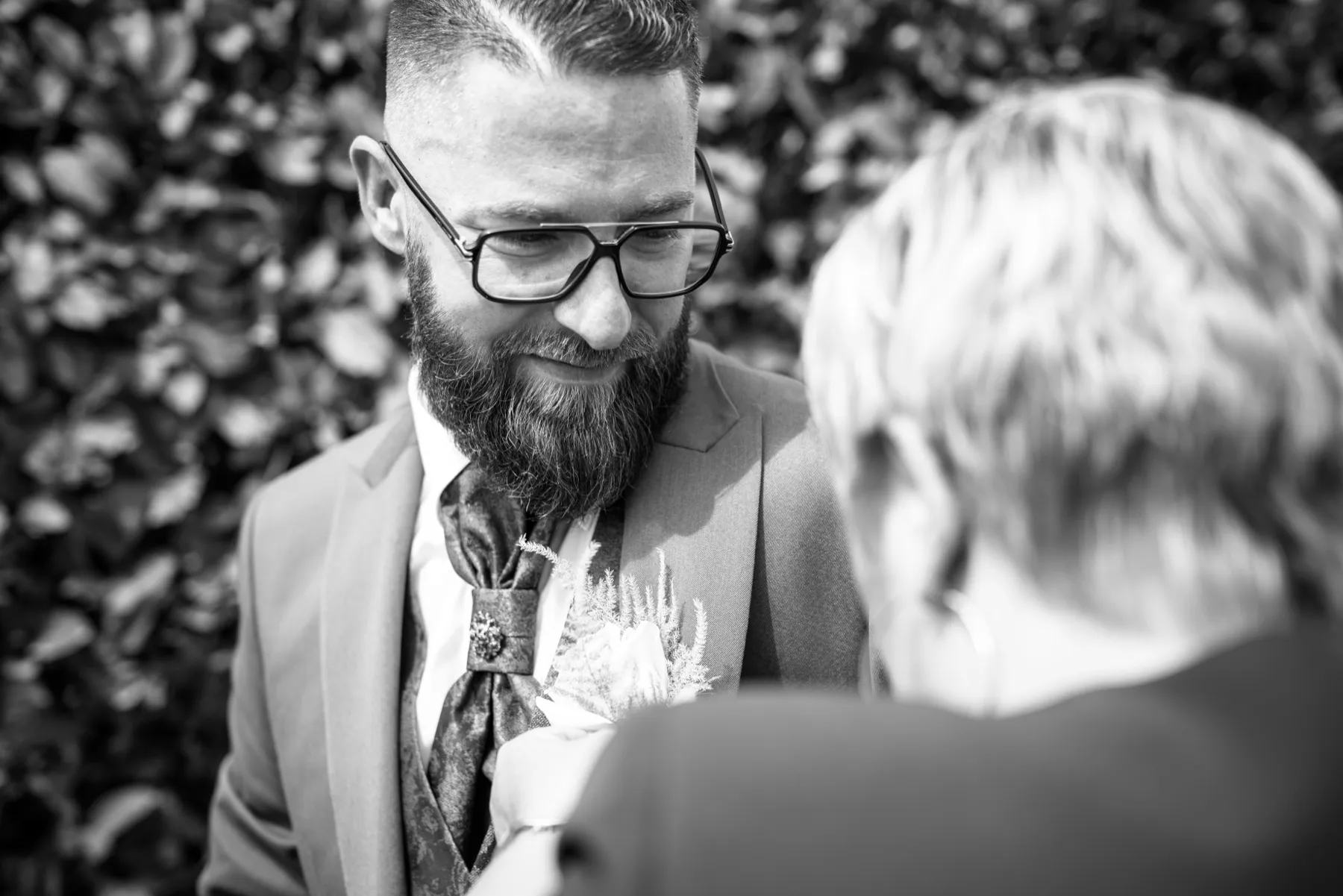 En noir et blanc, une personne épingle la boutonnière du marié, qui baisse les yeux