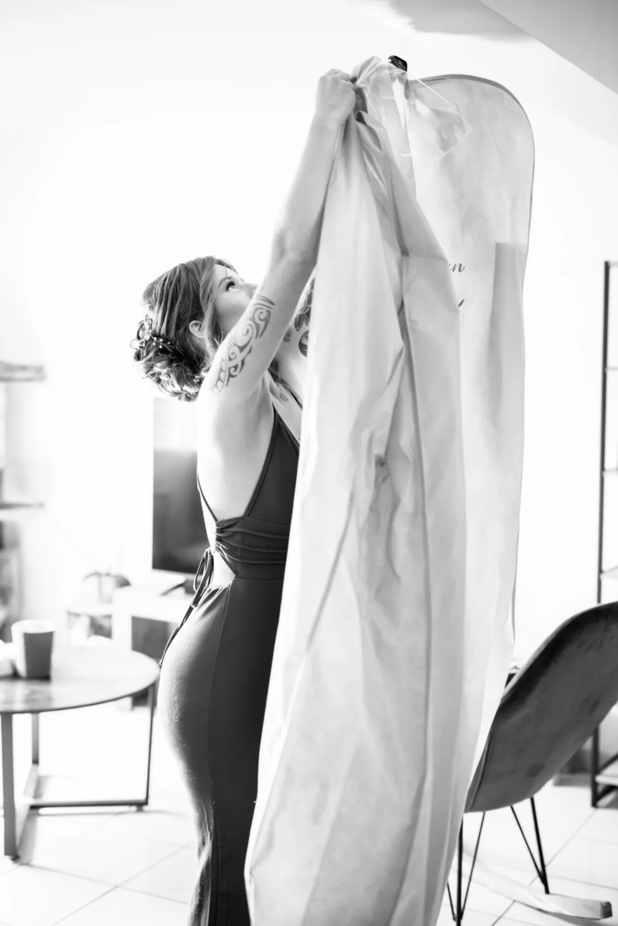 En noir et blanc, la future mariée soulève la housse qui protège sa robe