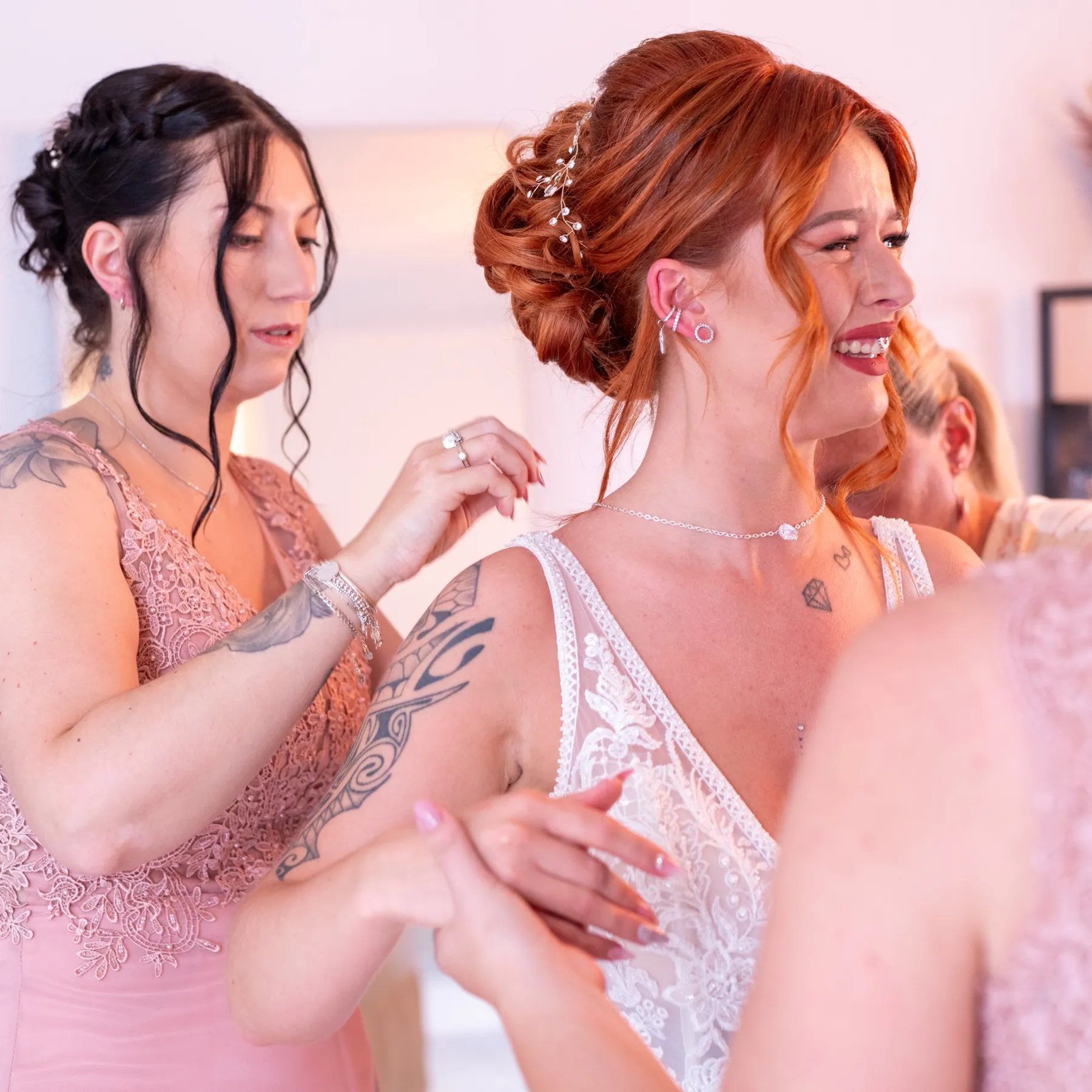 La mariée rit pendant qu’une proche en robe rose ajuste la bretelle de sa robe en dentelle
