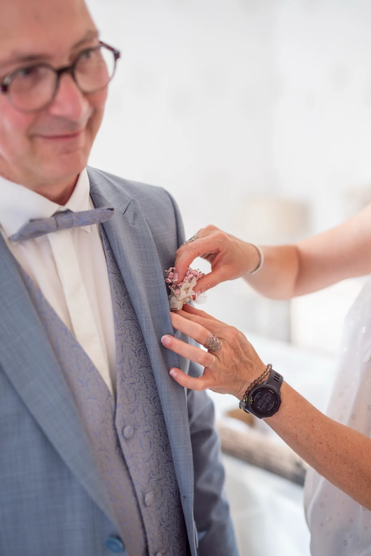 Une main épingle une fleur à la veste bleu-gris du papa de la mariée, qui sourit