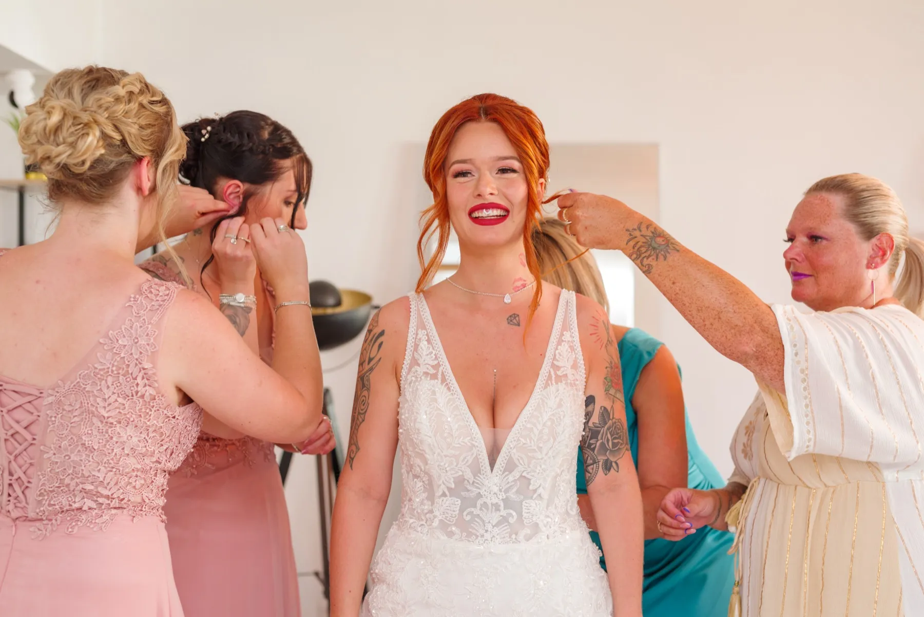La mariée sourit, en robe de dentelle, pendant que ses proches attachent ses boucles d’oreilles