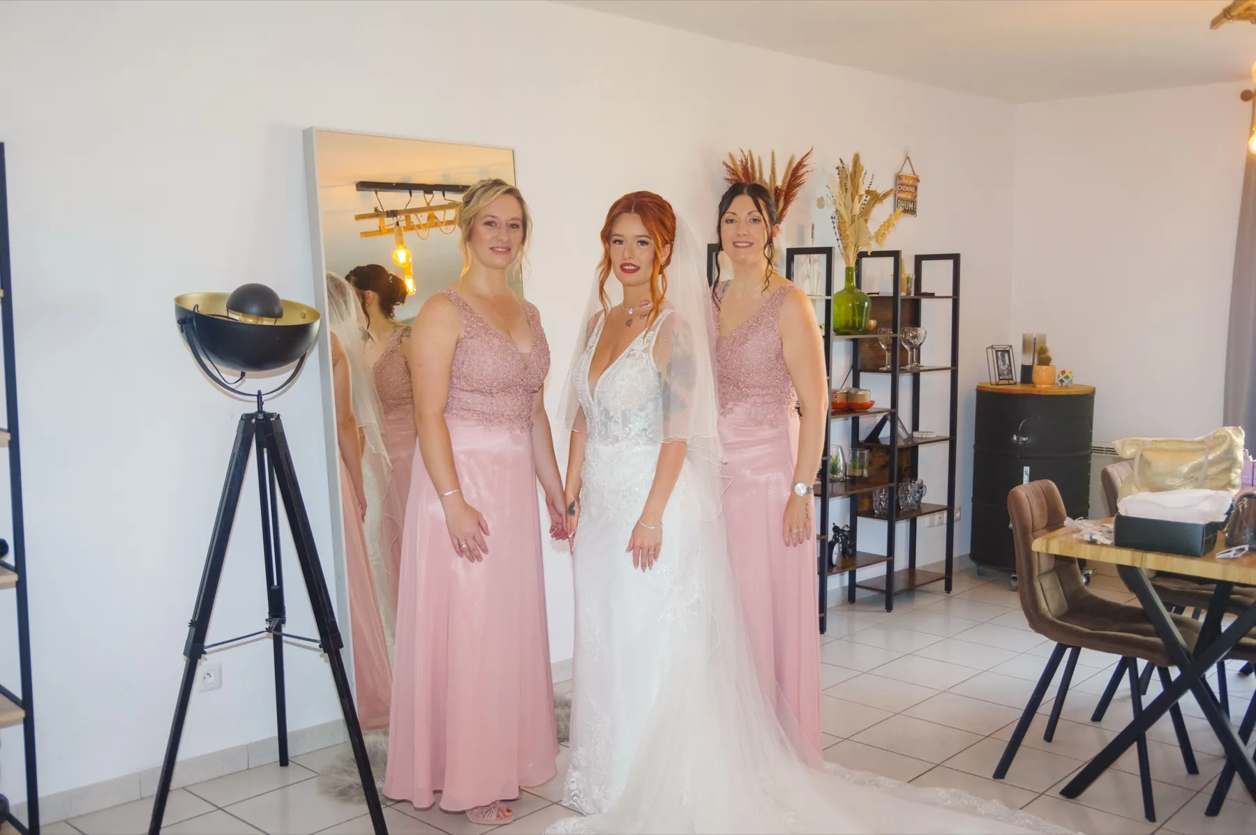 La mariée en robe et voile, entre deux proches en robe rose, dans le salon