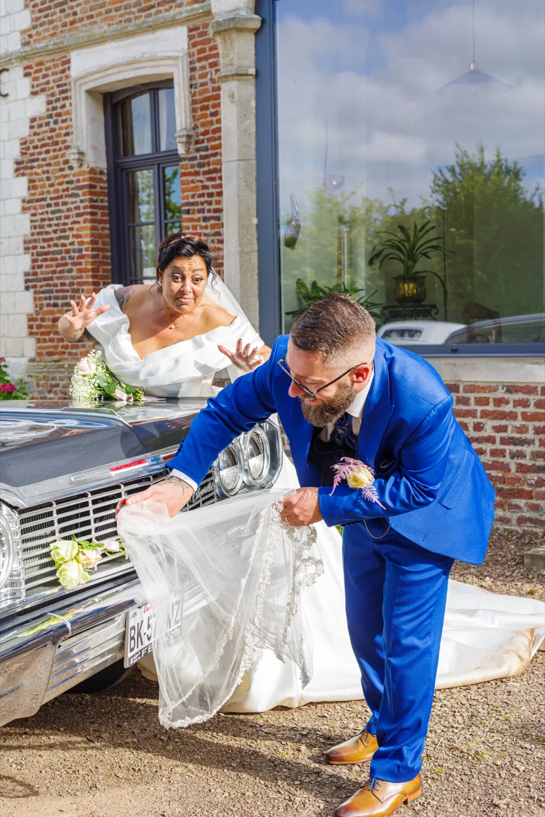 Le marié relève la traîne de la mariée qui fait une grimace comique près d'une voiture ancienne