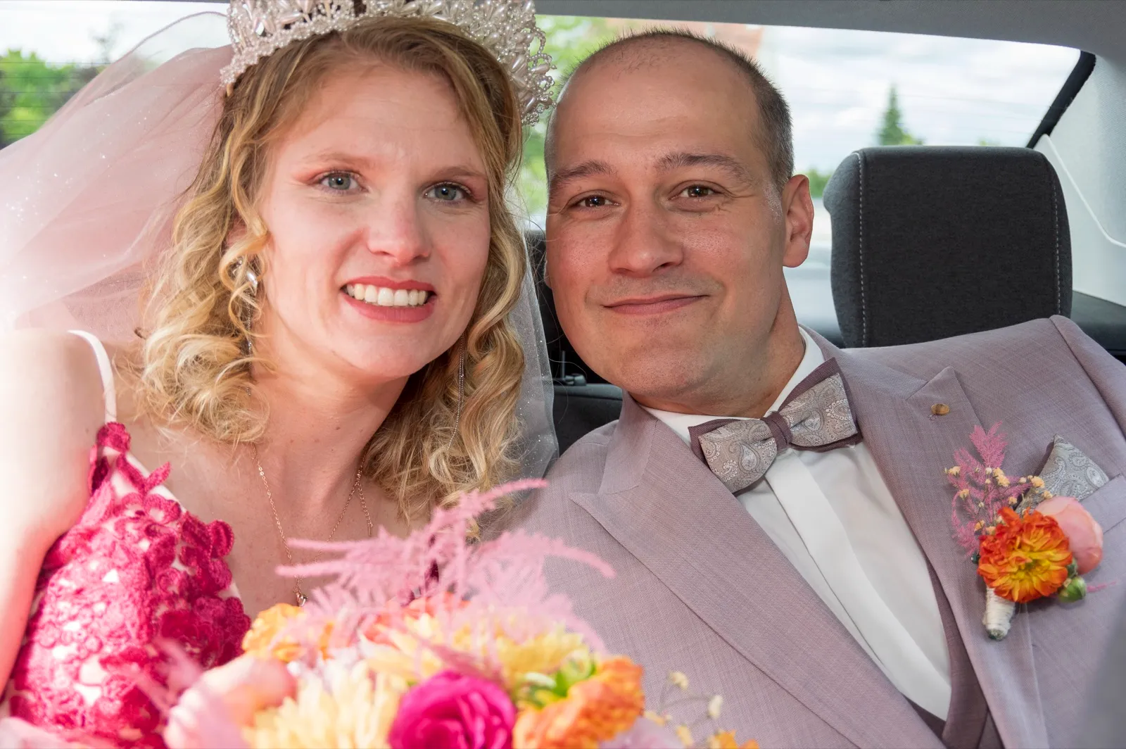 Dans une voiture, les mariés sourient à l'objectif, la mariée coiffée d'un diadème, bouquet coloré devant