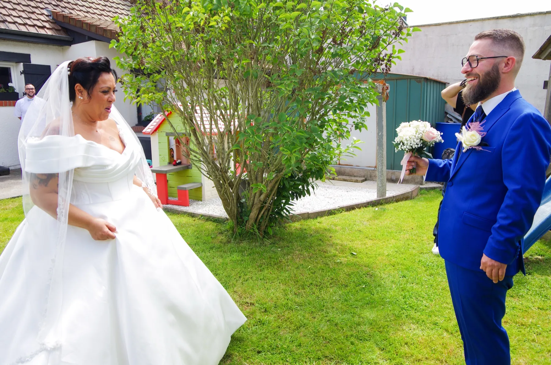 Dans le jardin, le marié, le bouquet à la main, rit en découvrant la mariée dans sa robe
