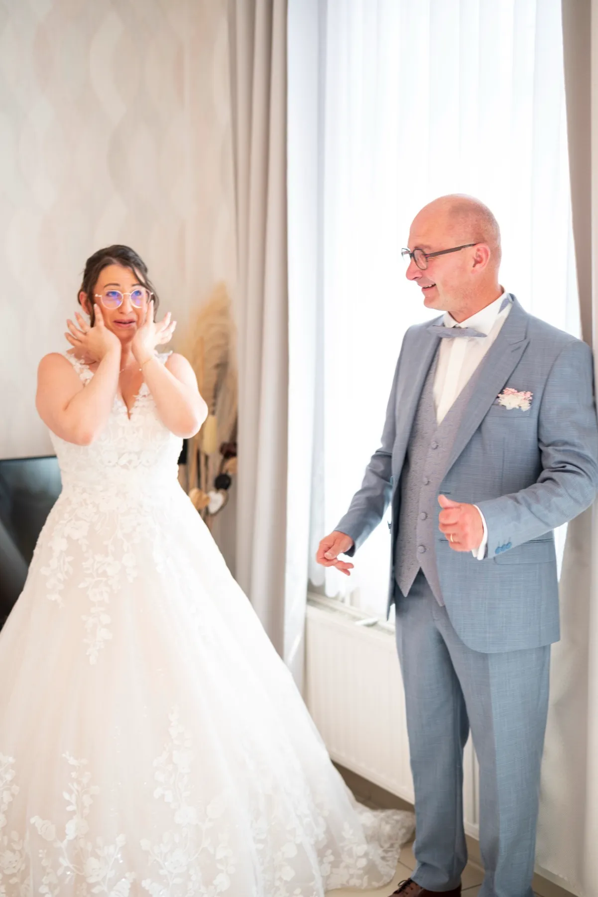 La mariée porte les mains à son visage ; son papa, en costume bleu-gris, la découvre dans sa robe