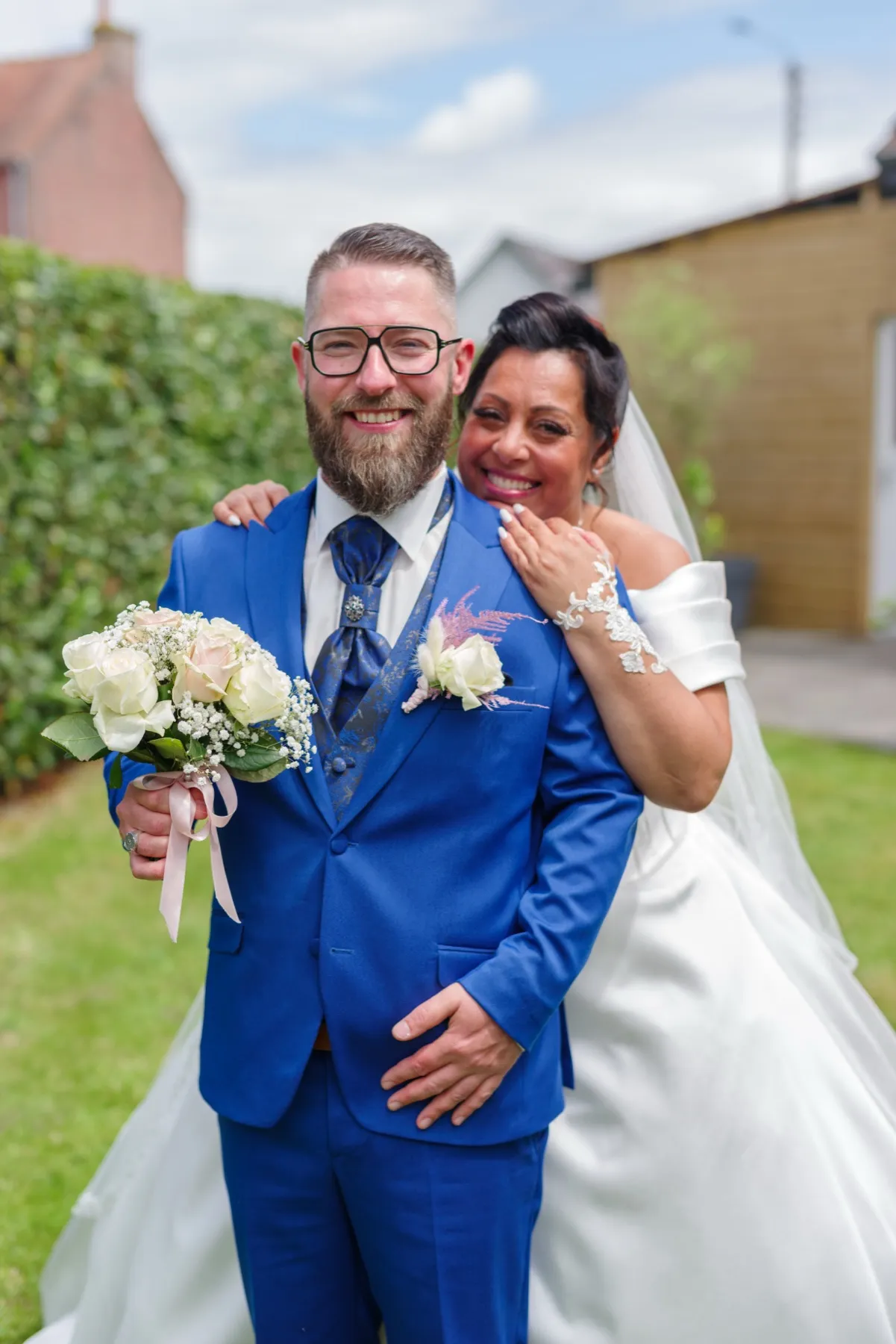 La mariée, rieuse, se glisse derrière le marié et pose la main sur son épaule