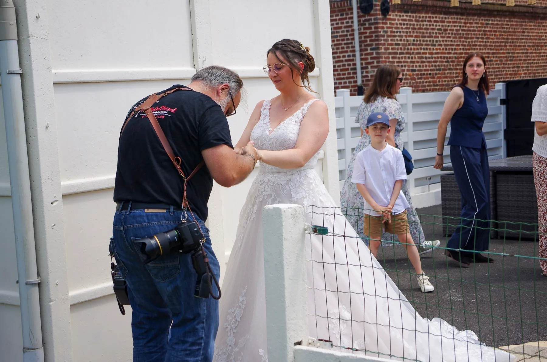 Vu de dos, le photographe guide la mariée vers la découverte, devant une barrière blanche