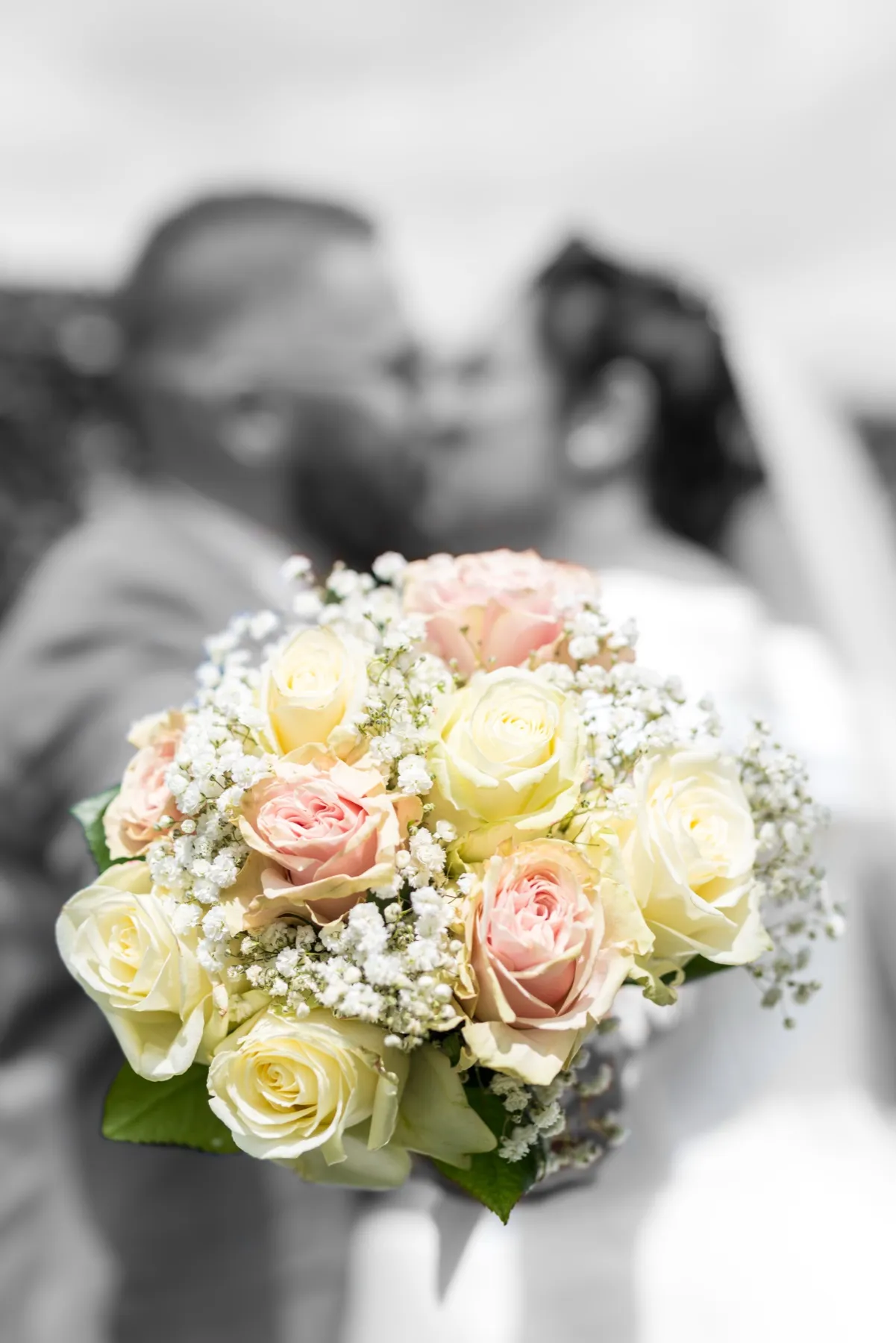 Le bouquet de roses pâles en couleur au premier plan, les mariés s’embrassent en noir et blanc