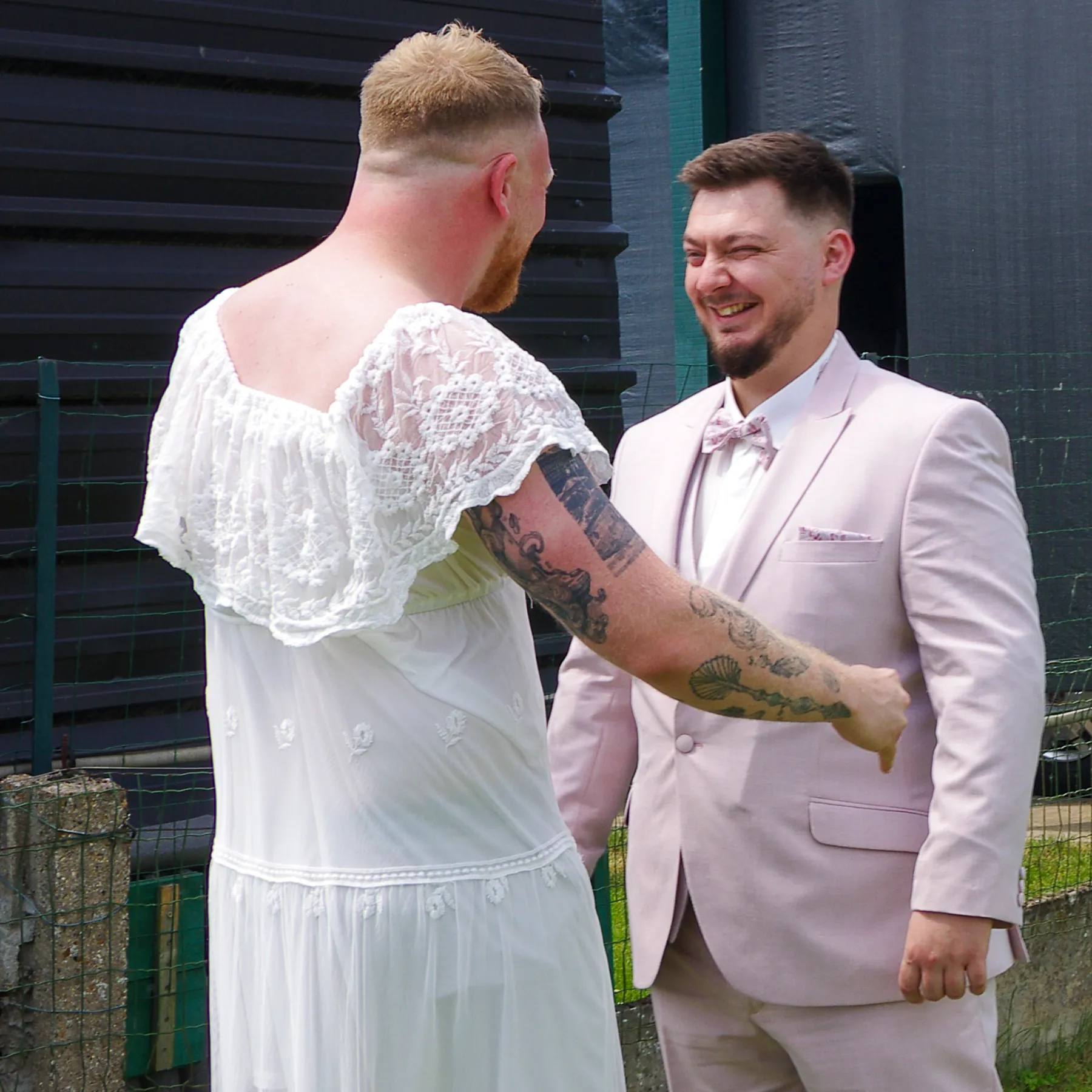 Le marié en costume rose pâle éclate de rire face à son meilleur copain, vêtu d’une robe de dentelle