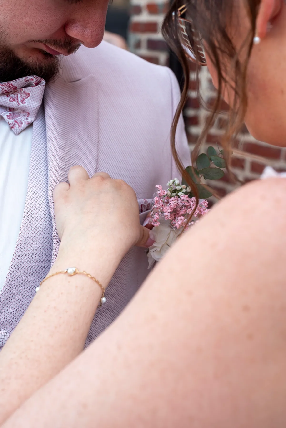 La mariée épingle la boutonnière fleurie du marié en costume rose pâle