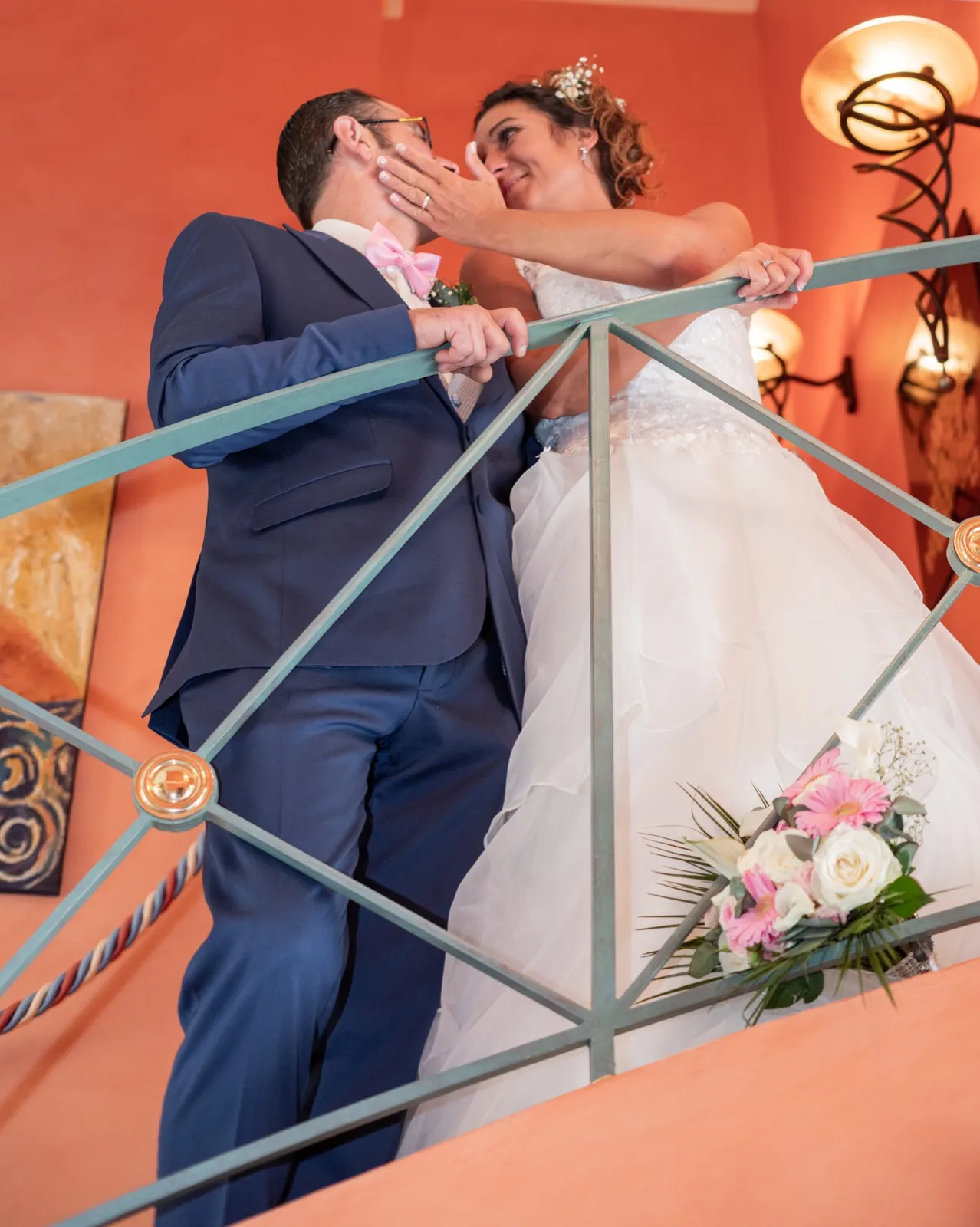 Vus d'en bas dans un escalier orangé, les mariés s'embrassent près d'un bouquet rose