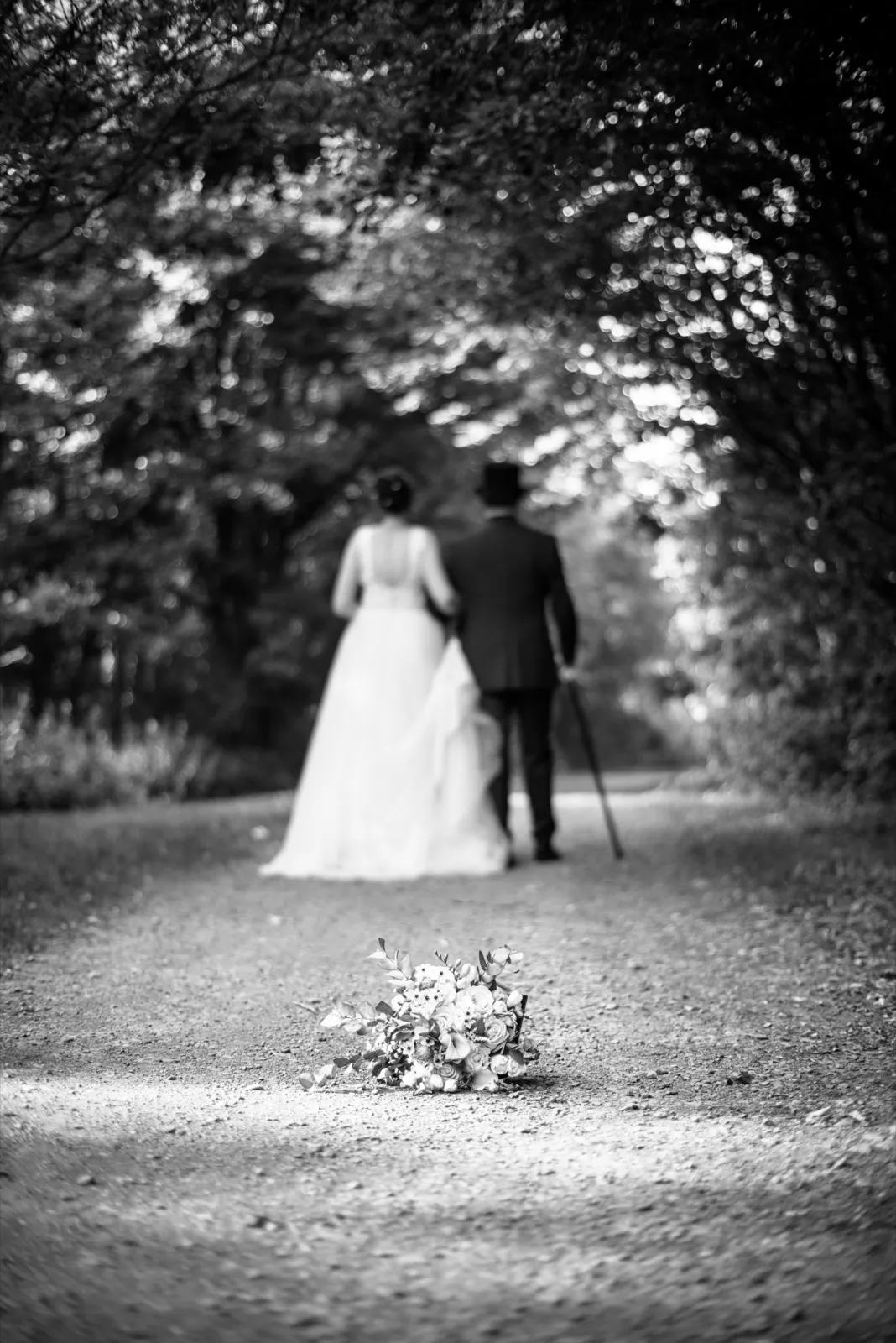 En noir et blanc, les mariés s'éloignent sur un chemin, un bouquet posé au sol derrière eux