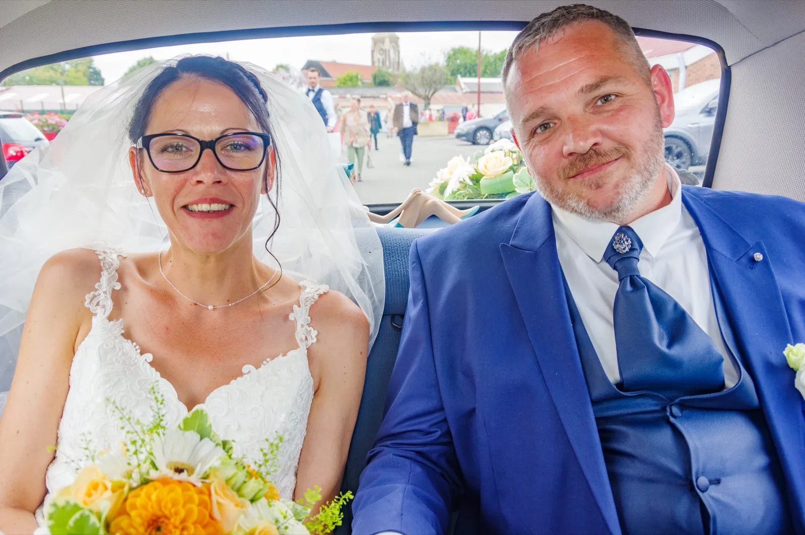 Assis dans une voiture, les mariés sourient à l'objectif, un bouquet jaune et orange au premier plan