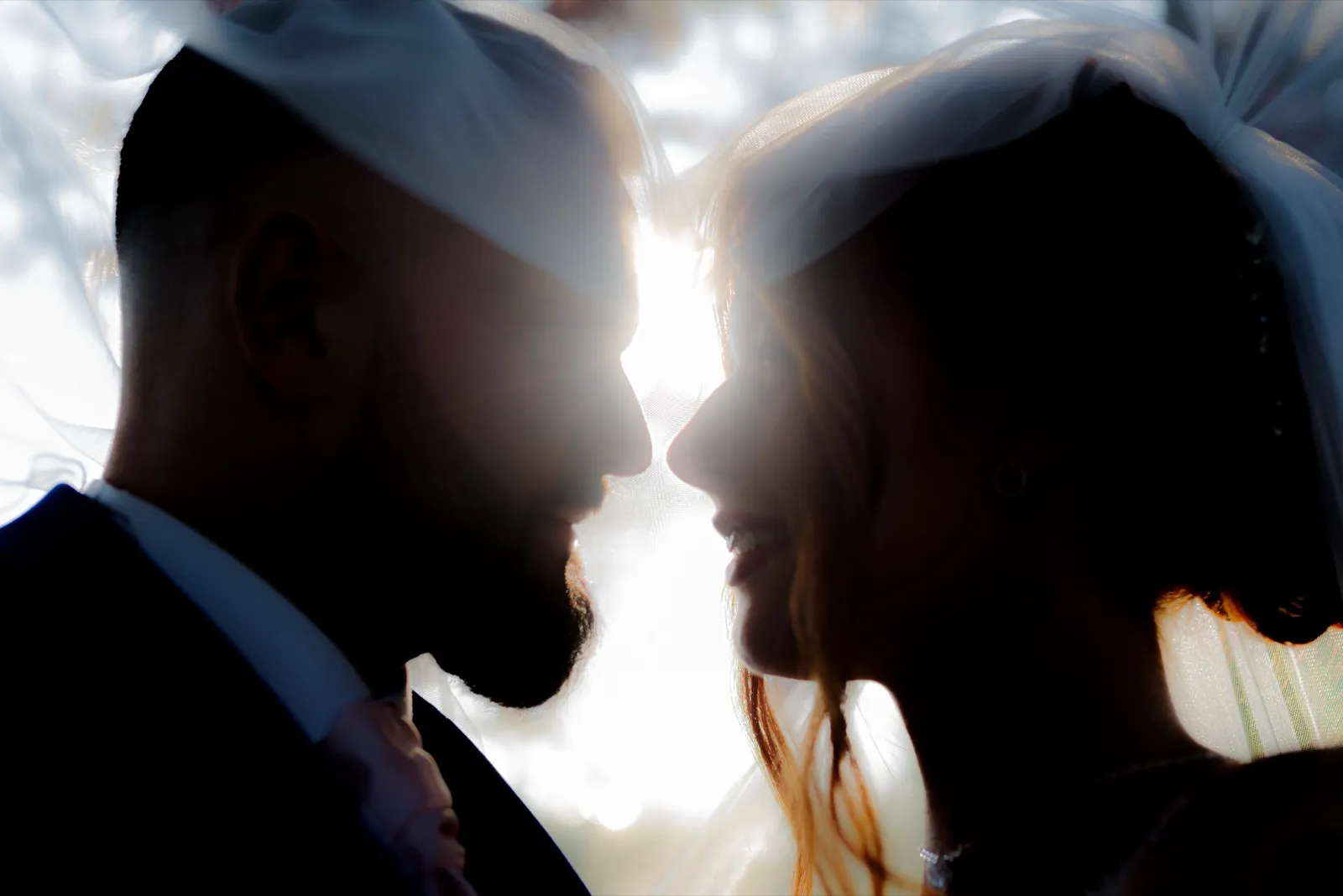 Les mariés se frôlent le nez sous un voile, en silhouettes à contre-jour