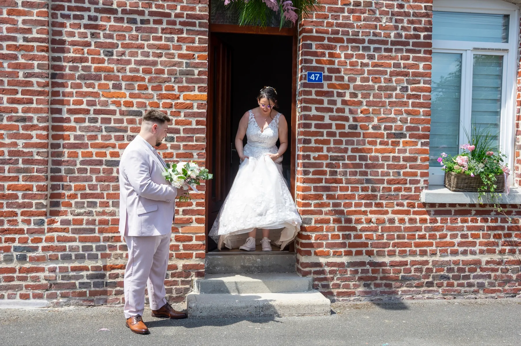 La mariée sort d’une maison en briques, le marié l’attend sur le trottoir avec le bouquet