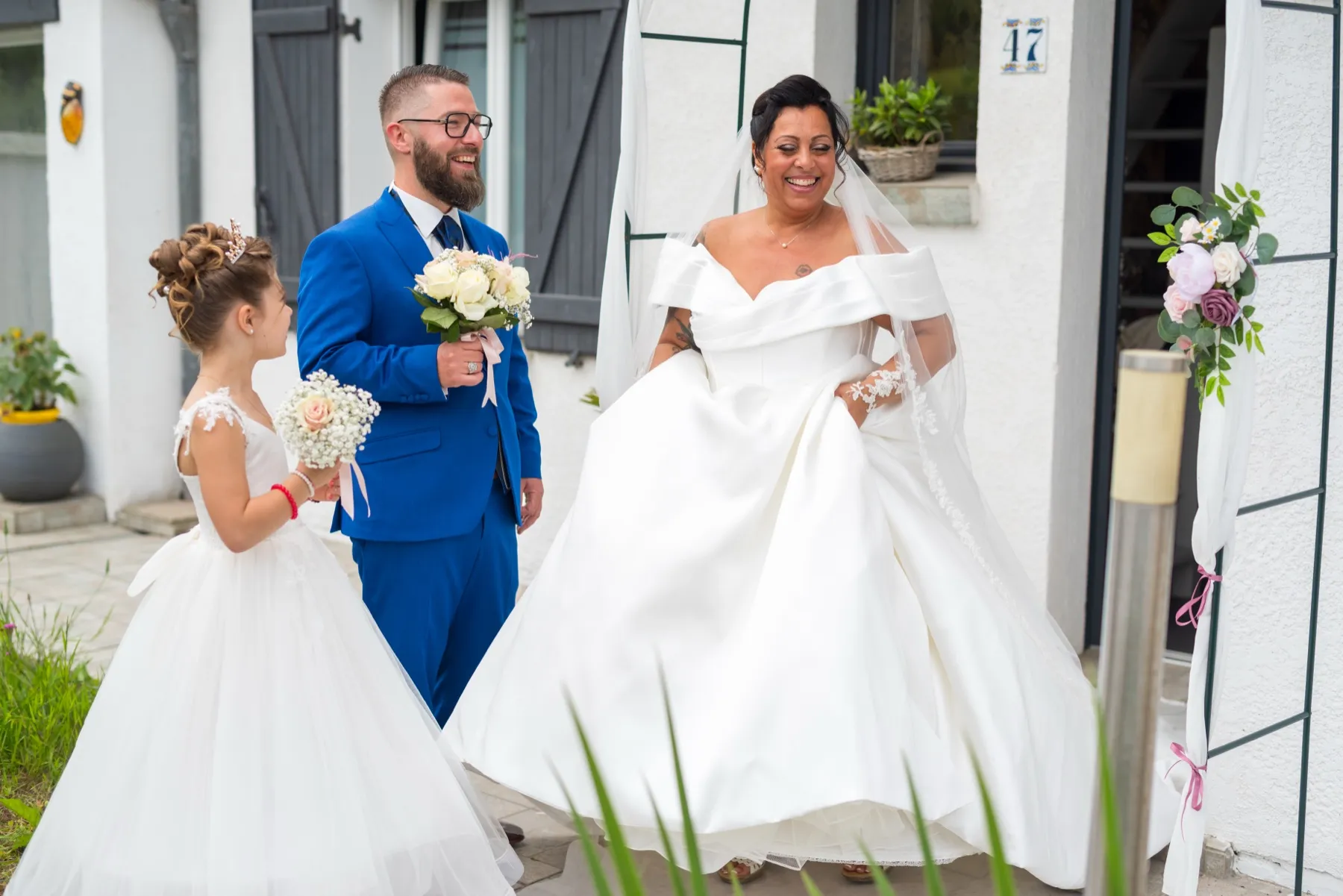 Devant la maison, le marié et une petite fille en robe blanche regardent la mariée s’avancer en riant