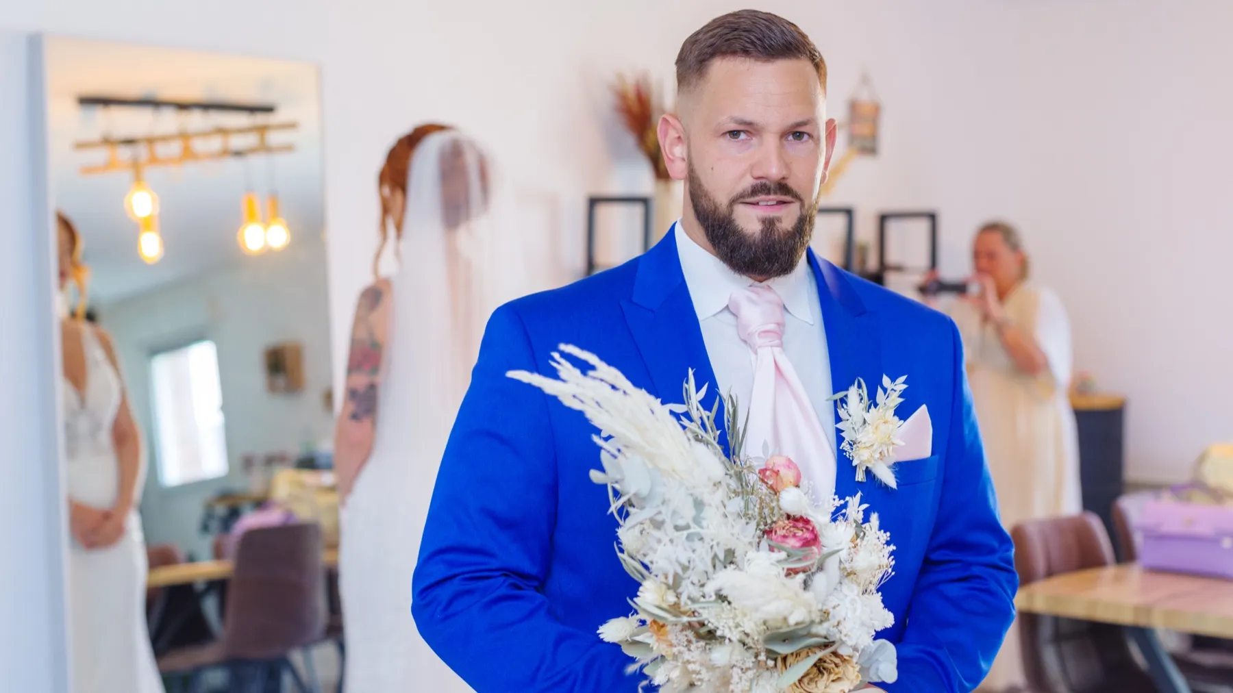 Le marié en costume bleu roi tient le bouquet, la mariée se reflète dans le miroir derrière lui