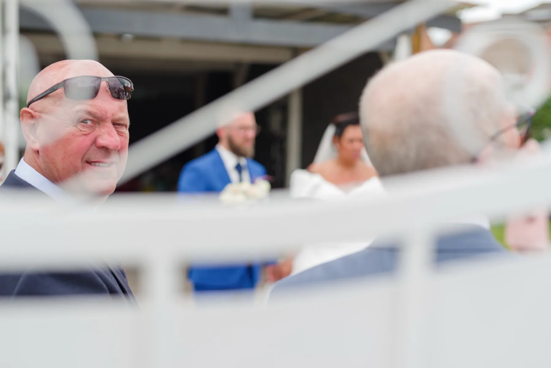 Derrière une barrière blanche, les mariés aperçus entre deux invités au premier plan