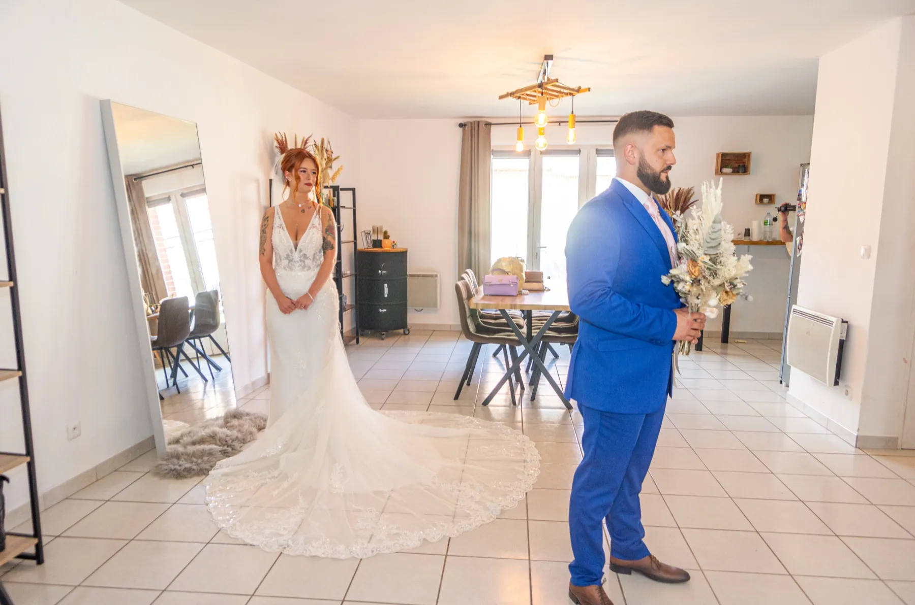 Le marié fait face à la mariée dans le salon, la traîne de dentelle étalée sur le carrelage