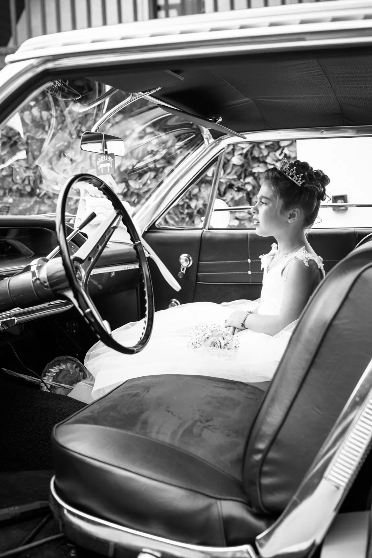 En noir et blanc, une petite fille en diadème assise à l’avant d’une voiture ancienne