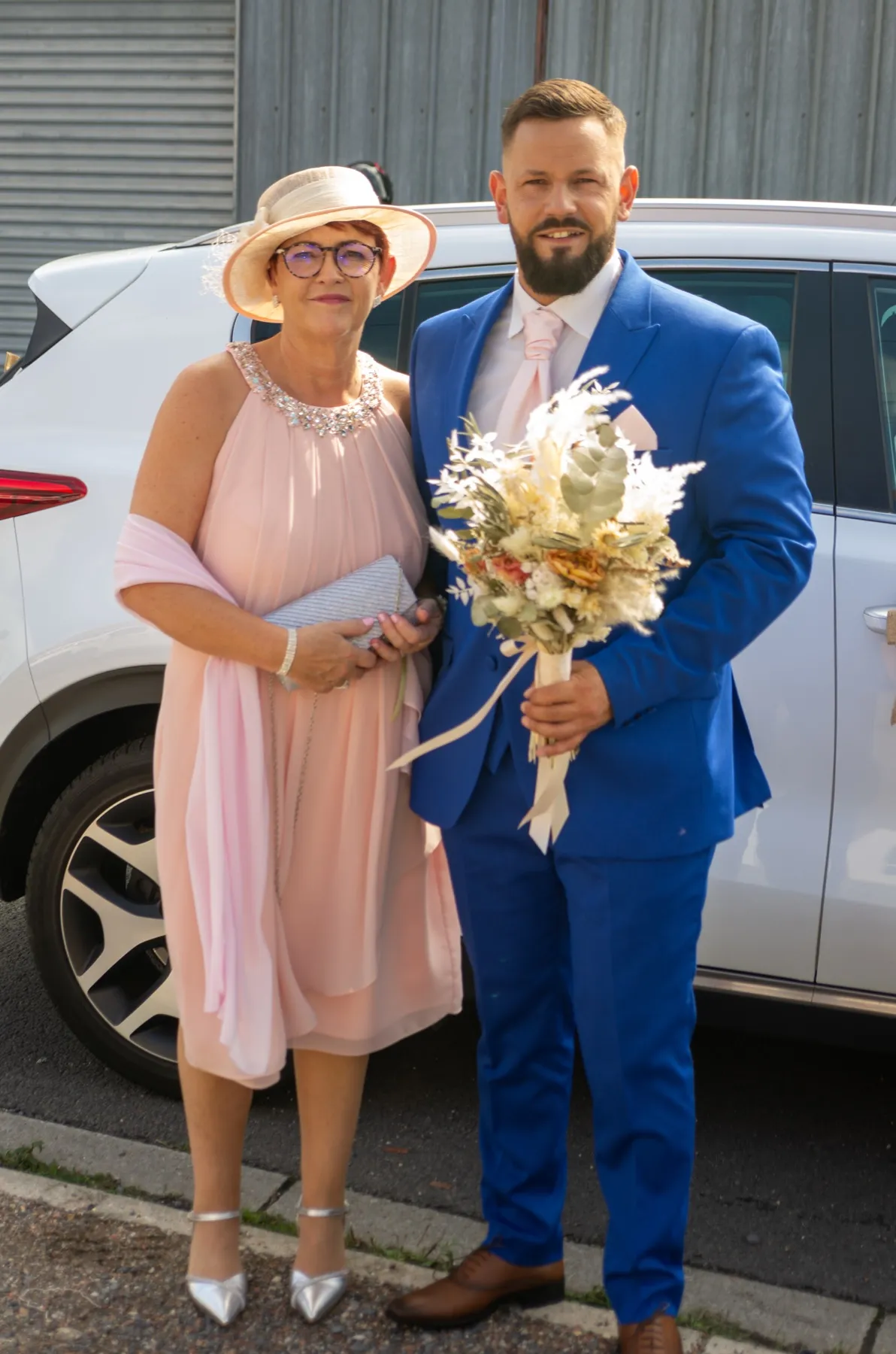 Le marié, un bouquet de fleurs séchées à la main, pose près d’une voiture avec une invitée en chapeau