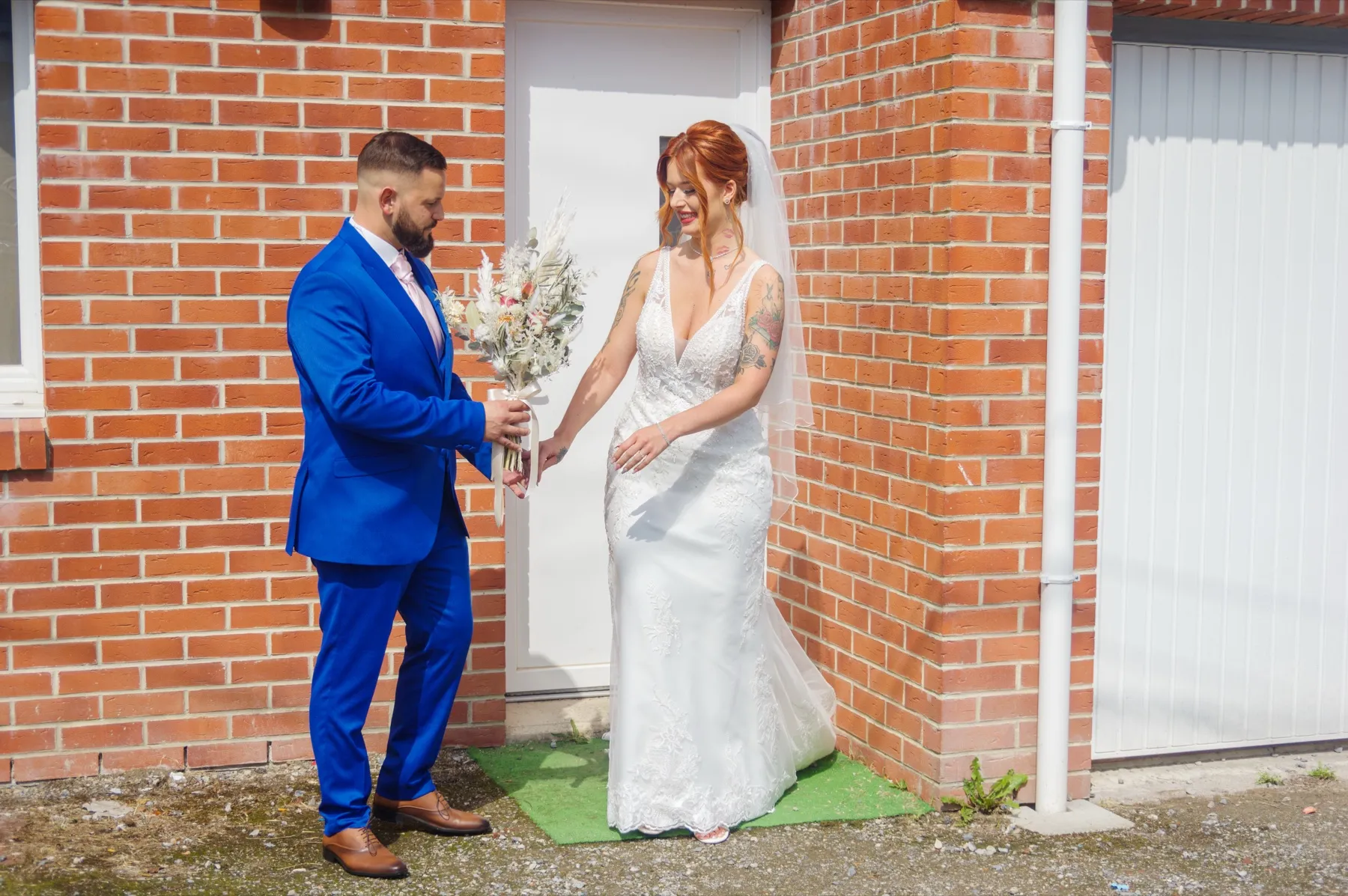 Devant la maison en briques, le marié tend le bouquet à la mariée