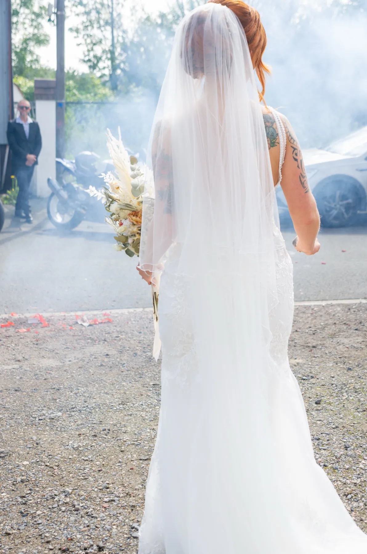La mariée de dos, sous son voile, s’avance dans la cour, le bouquet à la main
