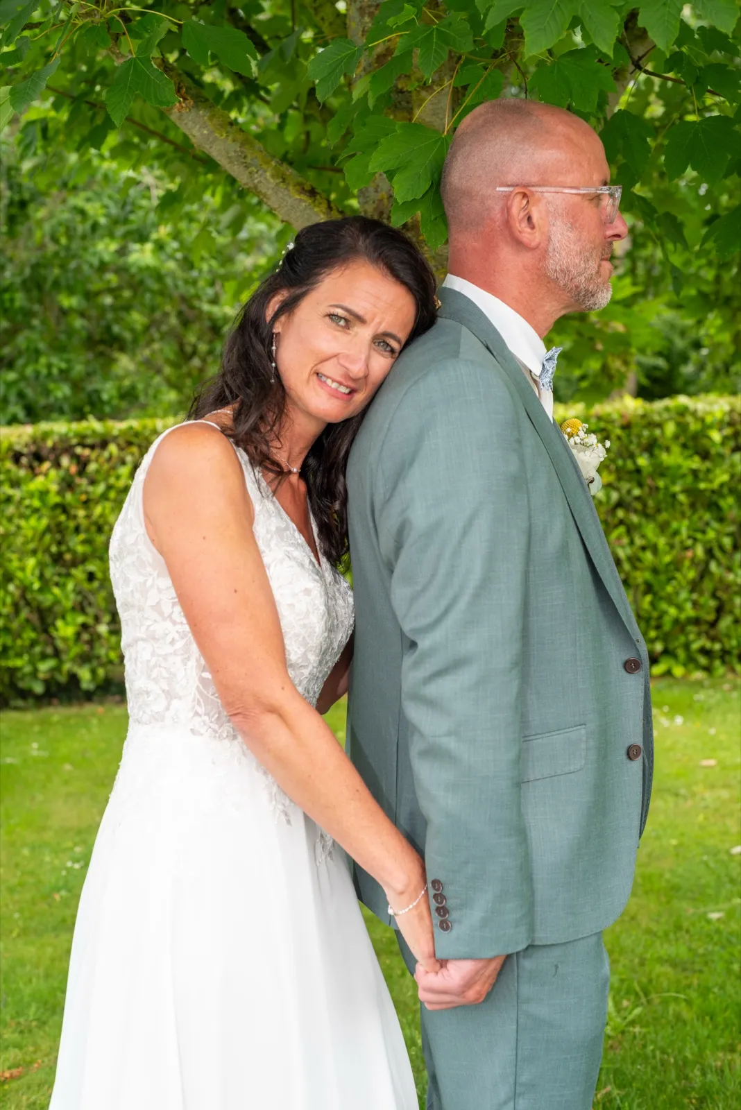La mariée appuie sa tête contre le dos du marié et sourit, devant une haie et un arbre