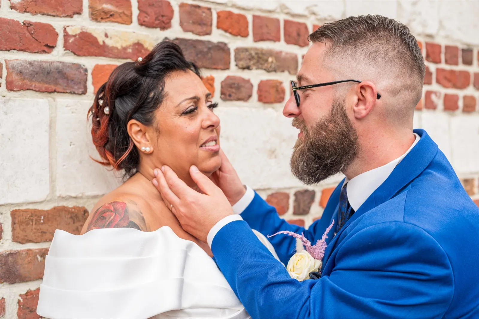 Le marié tient le menton de la mariée contre un mur de briques, tous deux souriants