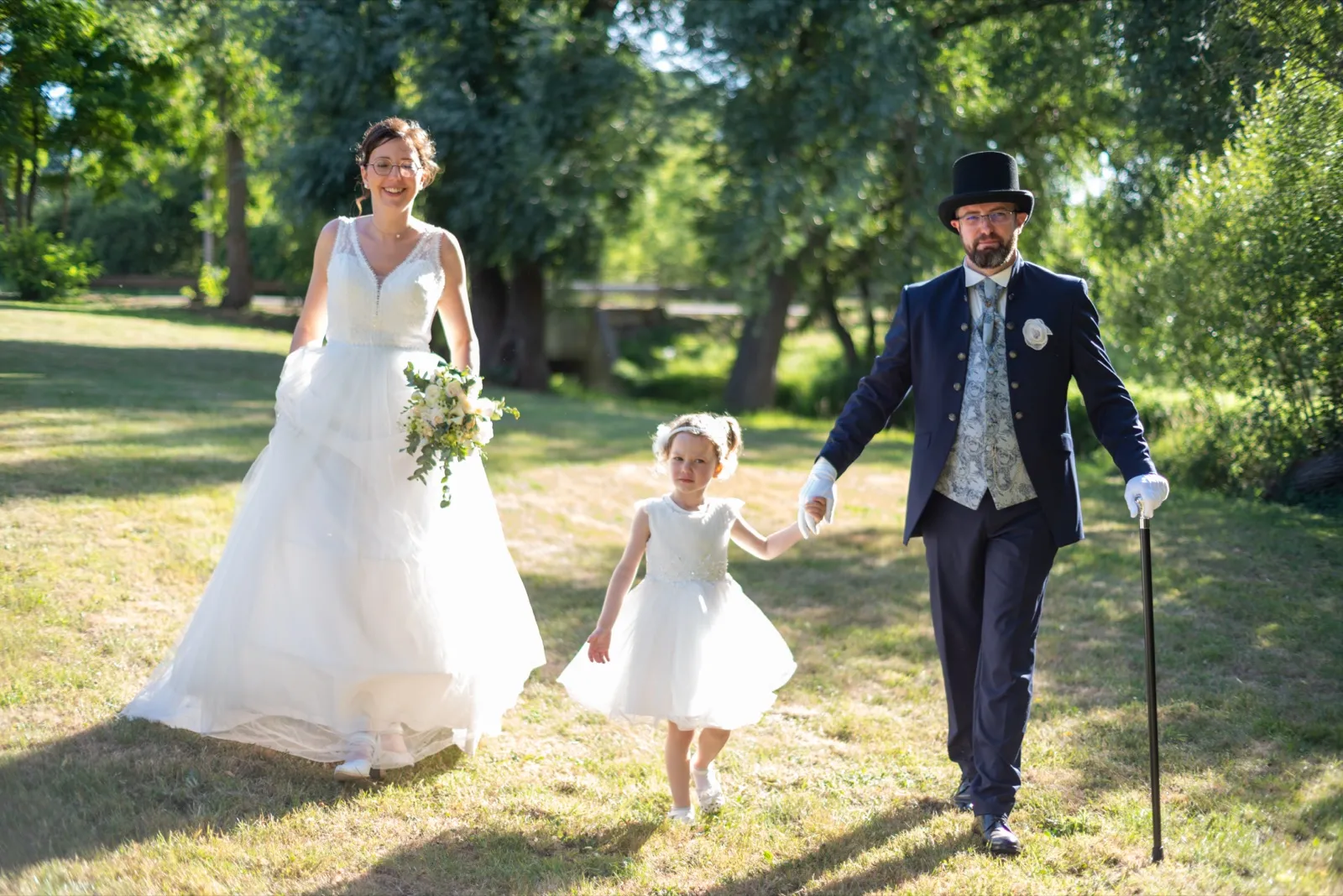 La mariée tenant son bouquet et le marié en haut-de-forme marchent avec une petite fille dans l'herbe