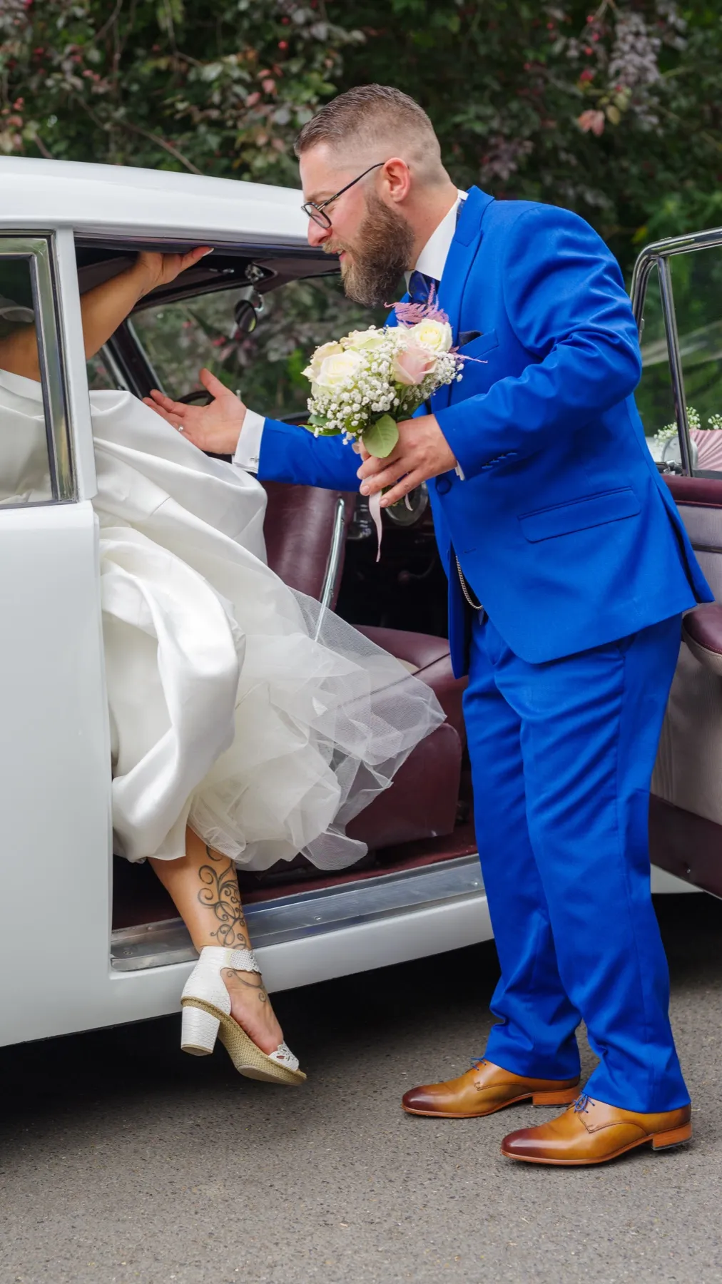 Le marié, le bouquet à la main, aide la mariée à descendre d’une voiture ancienne blanche