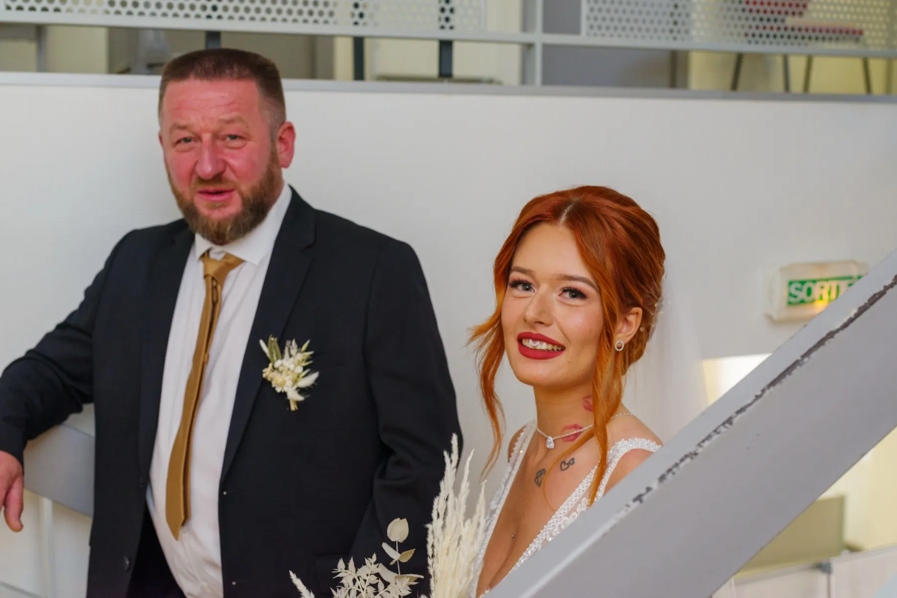 La mariée, souriante, près d’un escalier, à côté d’un homme en costume sombre