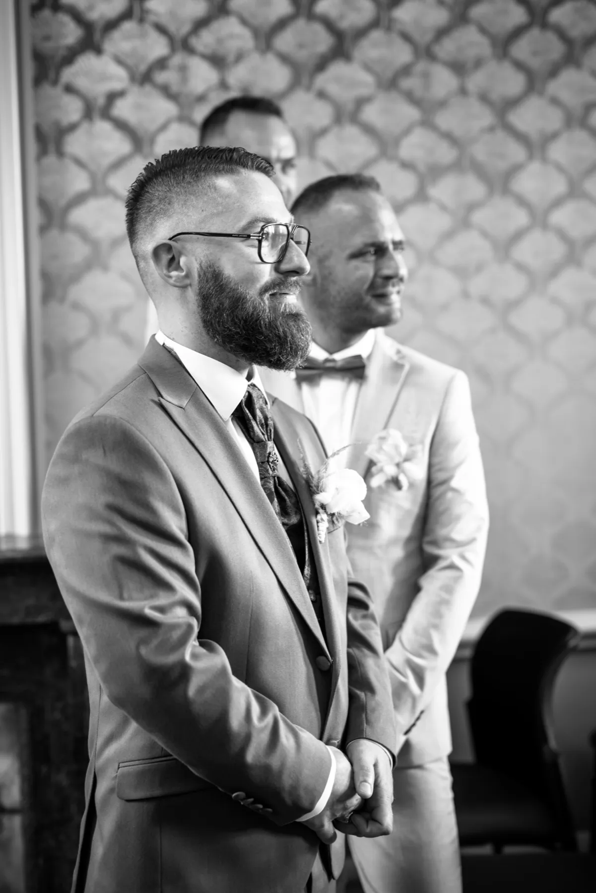 En noir et blanc, le marié debout attend en souriant, un homme en costume clair à ses côtés